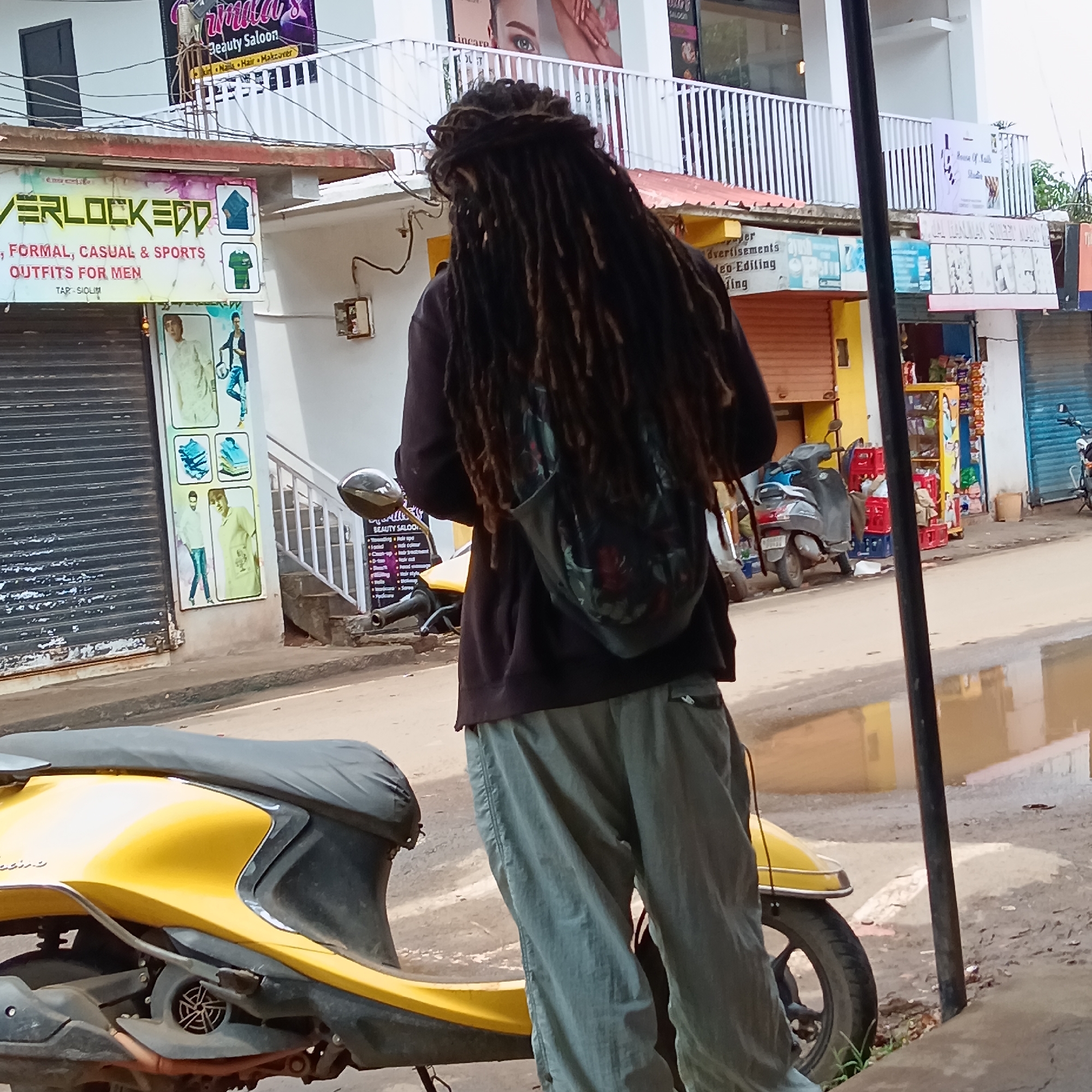 The image shows a street scene in Oxel, India. A person with long dreadlocks, wearing a dark hoodie and light-colored pants, stands facing away from the viewer, next to a yellow motor scooter. The person has a dark backpack. Behind the person are buildings with storefronts. One store is labeled "VERLOCKEDD, FORMAL, CASUAL & SPORTS OUTFITS FOR MEN." Above it is a sign for "Rinaca's Beauty Saloon." Another shop is labeled "HANUMAN STEEL WORKS." The street is wet with puddles. The image captures a mundane urban environment.