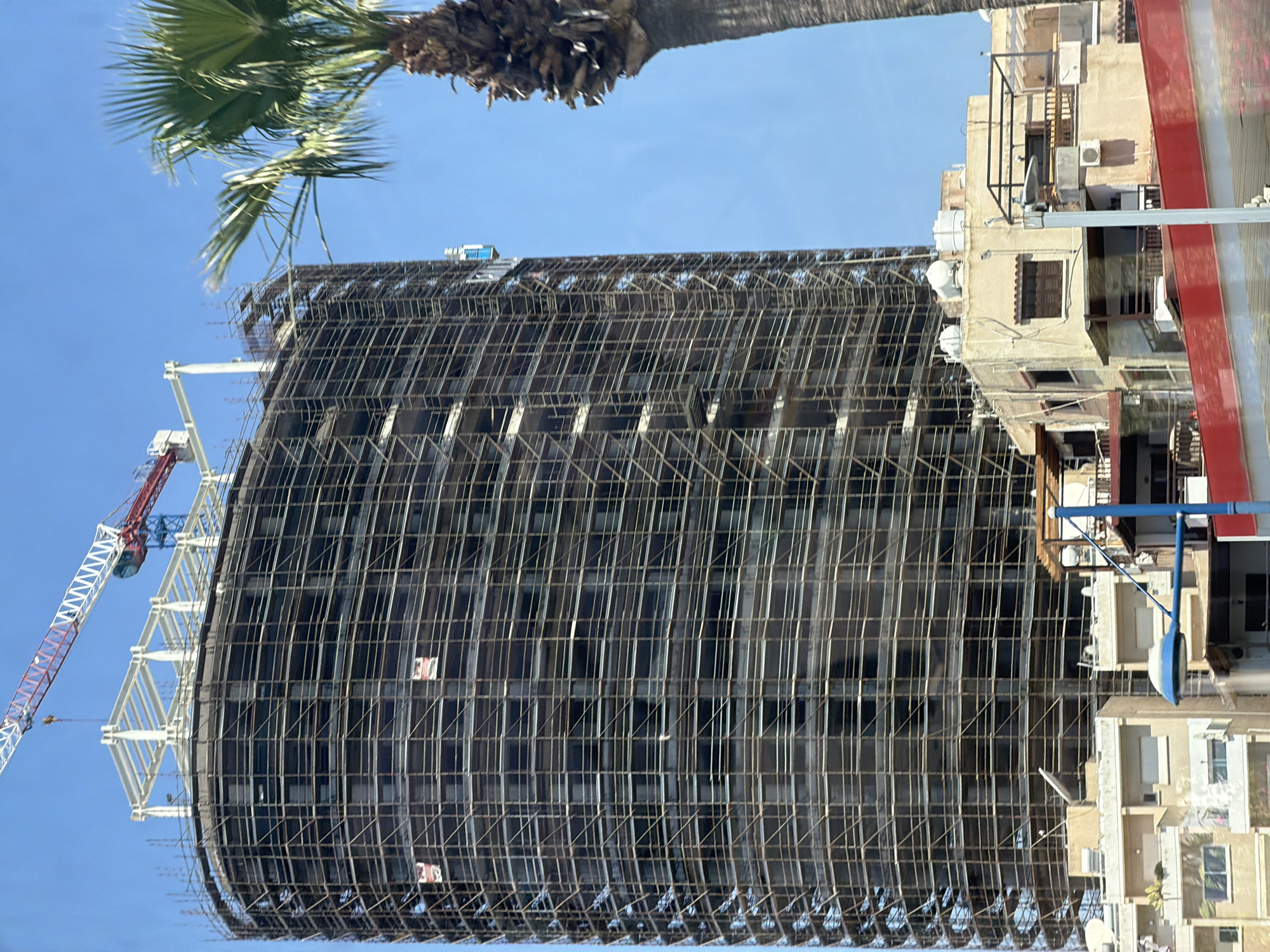 The image depicts a building under construction in Agios Tychonas Community, Cyprus. The building is surrounded by scaffolding. A construction crane is visible to the left of the building. Adjacent to the building are older structures. The sky is clear and blue. A palm tree is visible in the upper left corner of the image.
