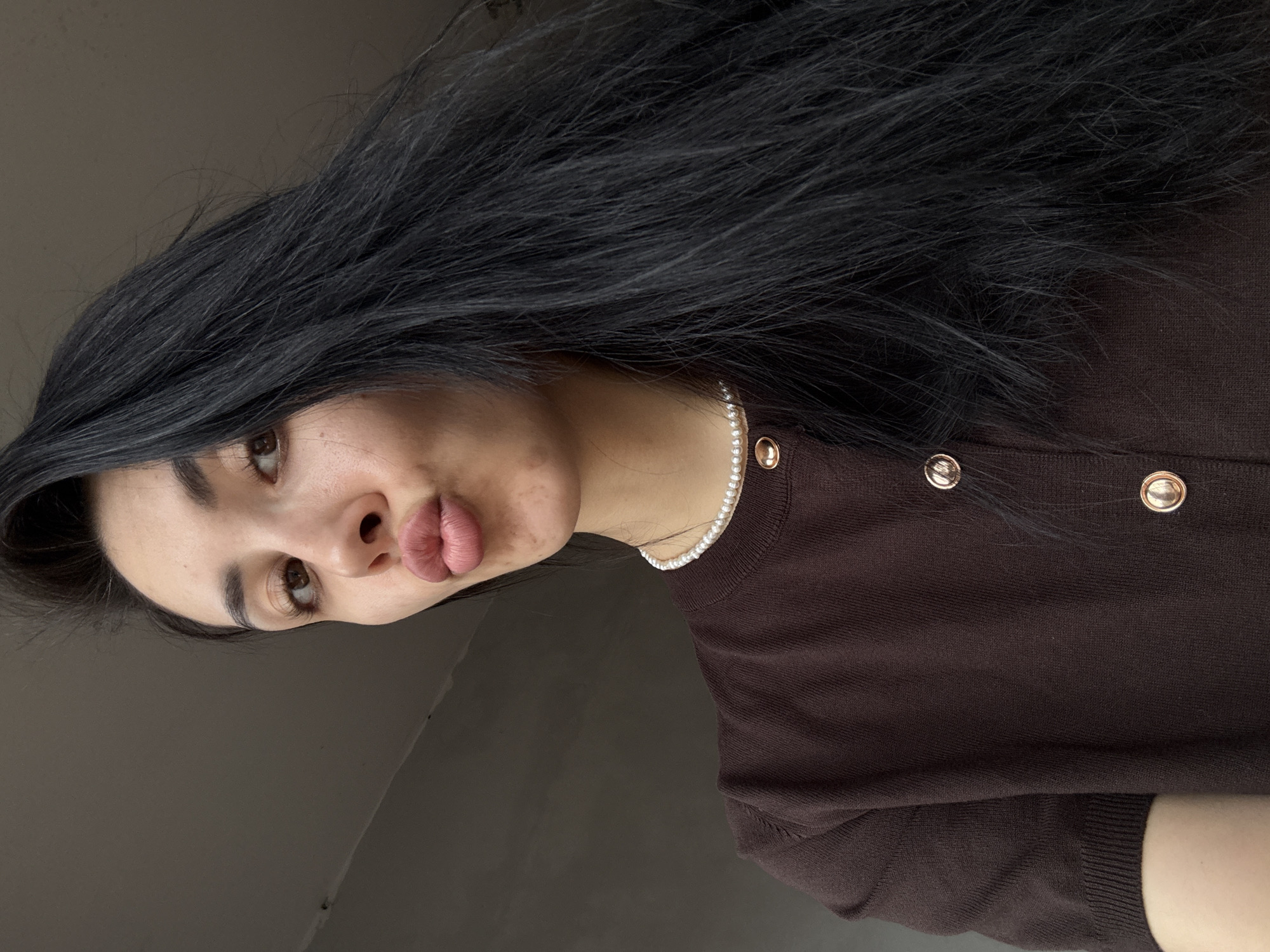 The image presents a close-up, angled portrait of a young woman with long, dark hair. Her face is turned slightly to the right and tilted upwards, with her lips pursed in a "kissy face" or "duck face" expression, indicating a posed shot. Her right eye, visible in the upper left of her face, has a brown iris and well-defined eyelashes, looking towards the upper left corner of the frame. Her skin appears smooth, with some subtle texture or very light blemishes visible on her cheek.
Her long, dark, and seemingly thick hair sweeps across the top and left side of the image, appearing slightly windswept or styled to fall naturally. She is wearing a dark brown top, possibly a finely knitted cardigan or sweater, featuring several round, shiny, gold-toned buttons down the front. A delicate pearl necklace adorns her neck, adding a touch of elegance.
The background is a plain, solid dark gray or brown surface, suggesting an indoor setting without any specific decor or environmental features. The lighting is soft and even, highlighting her features without creating harsh shadows. No specific activity beyond posing for the photograph is taking place. There are no visible texts or specific location cues within the image itself to suggest a particular time of day or the given context of Votkinsk, Russia. The overall impression is a casual, intimate portrait focusing on the subject's expression and details.