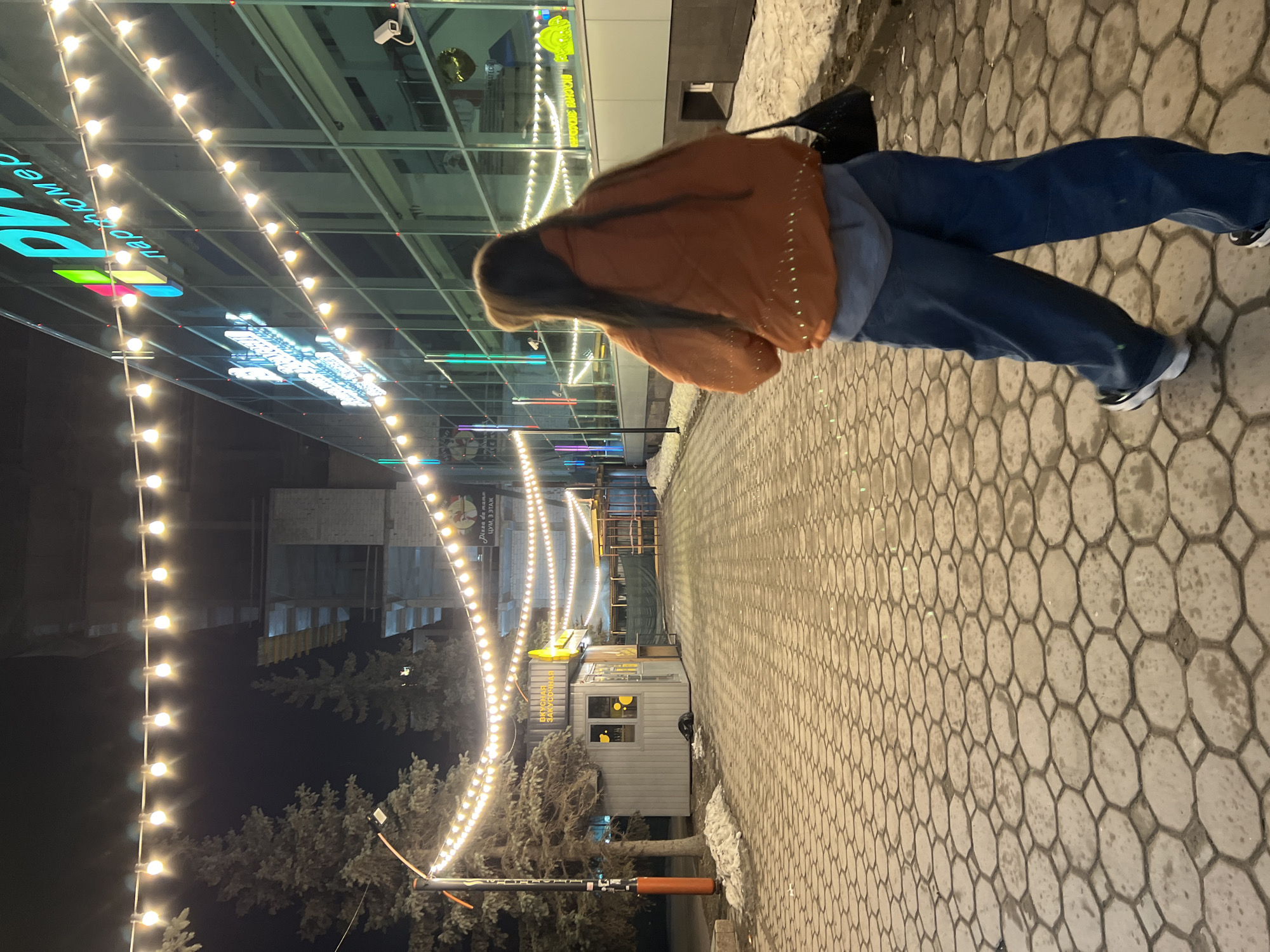 A photo taken in the city of Tyumen, Russia, at night, shows a young woman walking on a paved walkway. The walkway is made of cobblestone and is lined with streetlights. There is a building to the left of the walkway, and the walkway is lit by string lights. The woman is wearing a rust-colored jacket and jeans. She has long hair and is carrying a purse.