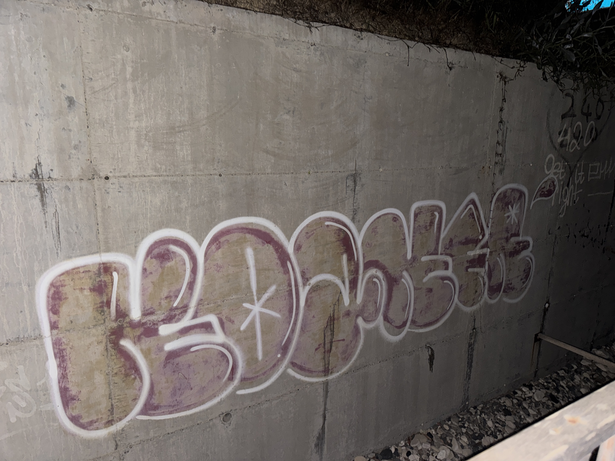 The image shows graffiti on a concrete wall in Agios Tychona Community, Cyprus. The graffiti consists of large, stylized letters in shades of brown and white, with a purple outline. The letters appear to spell out a word, but it is difficult to decipher completely due to the stylized nature of the graffiti. There are also some smaller markings on the wall, including numbers and what appears to be text, to the right of the main graffiti. The concrete wall is rough and weathered, with some cracks and stains. The area around the wall is covered in gravel and rocks, and there is some vegetation growing at the top of the wall. The lighting suggests it is nighttime or a dimly lit area.