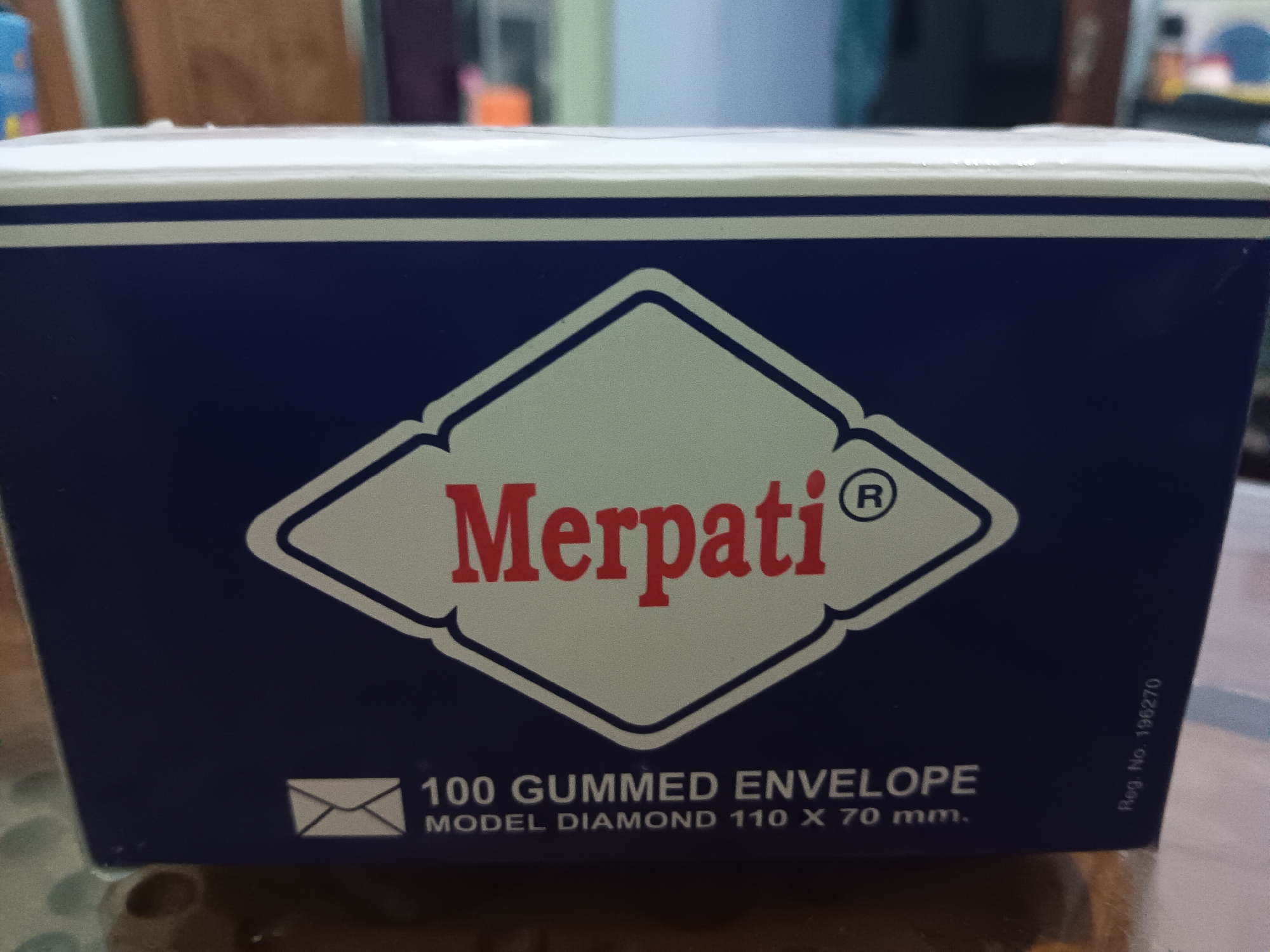 A close-up, eye-level shot shows the front of a dark blue cardboard box. The box has a white top edge with two horizontal blue stripes. Centered on the front of the box is a diamond shape outlined in blue, with a white interior. Inside the diamond, the word "Merpati" is written in red, stylized, sans-serif capital letters. To the right of "Merpati" is a registered trademark symbol (R) in a circle. Below the diamond and toward the bottom left of the box, there is a white icon of an envelope. To the right of the envelope icon, white text reads "100 GUMMED ENVELOPE" and below that "MODEL DIAMOND 110 X 70 mm." Along the right edge of the box, vertically oriented white text reads "Reg. No. 196270." The box is placed on a surface that appears to be a wooden table. The background is blurred and out of focus, with hints of vertical structures and possibly some colorful objects. The lighting suggests indoor ambient light. The image has a slight tilt.