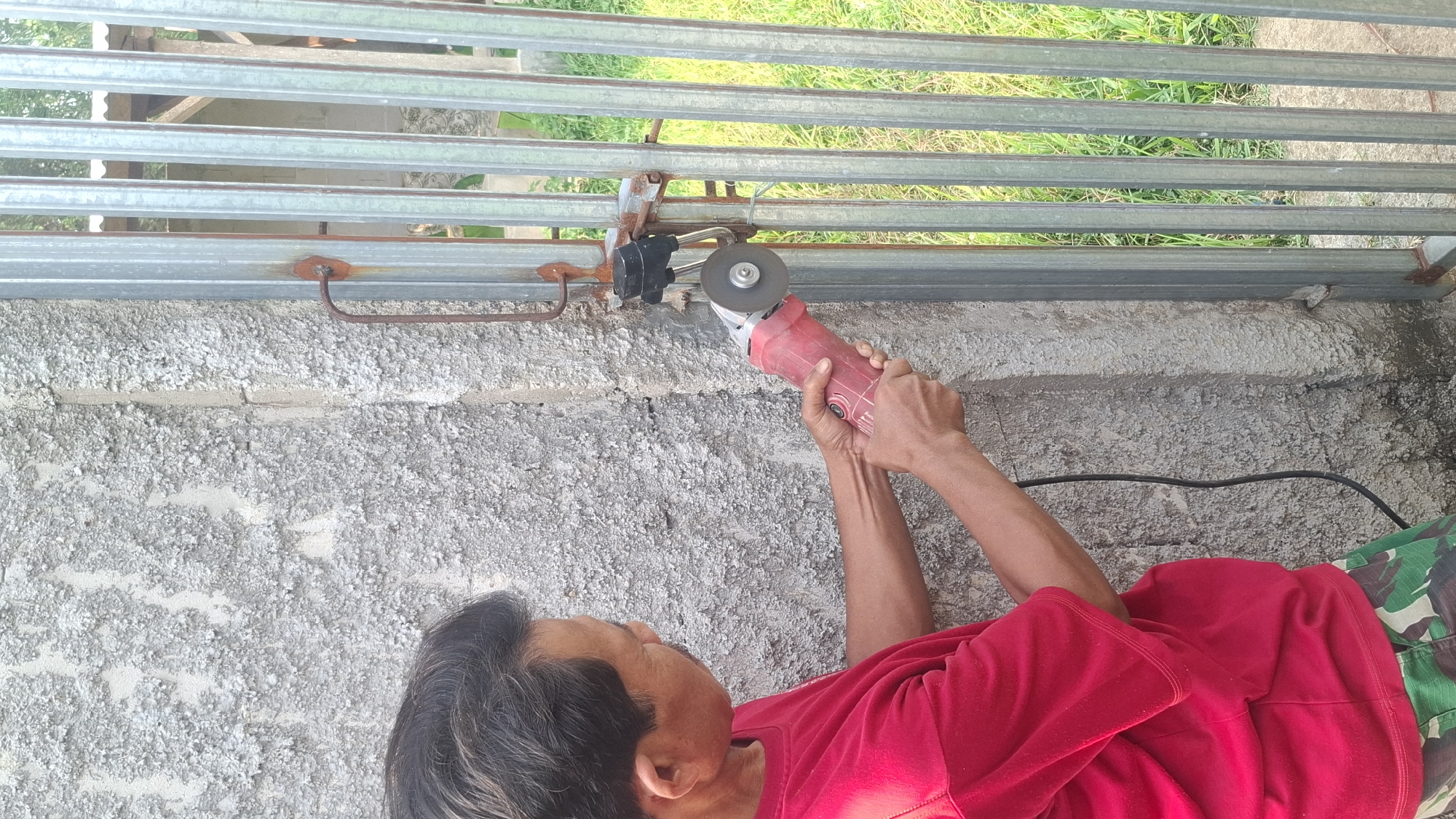 In Rahong, Indonesia, a man wearing a red shirt and camouflage shorts is using a red and silver angle grinder to cut through a metal gate lock. The man is standing next to a textured gray concrete wall and a vertical metal gate. Green grass is visible in the background behind the gate.