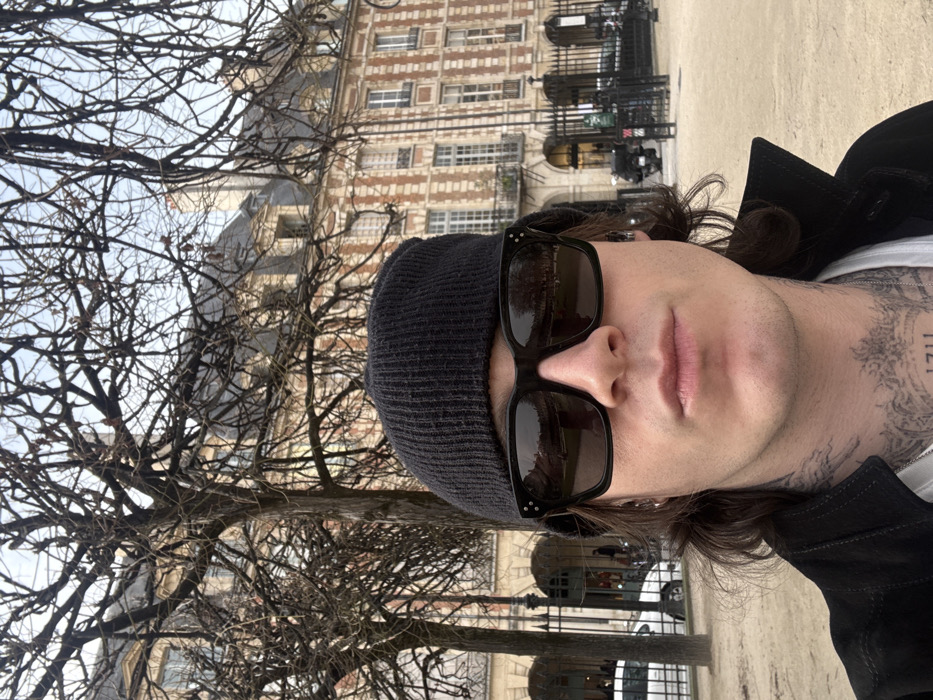 The image captures a male individual in a close-up selfie within the Place des Vosges in Paris, France. The subject, positioned in the foreground, wears a dark ribbed beanie and dark, rectangular sunglasses. His medium-length dark hair is visible below the beanie, and he has ear piercings. He is dressed in a dark, collared jacket over a lighter-colored shirt, with a silver chain visible. Prominent tattoos are visible on his neck, including the numbers "1711" below a crown-like design on his throat. In the midground, several bare deciduous trees with intricate branch networks are visible. The background features the distinctive architecture of the Place des Vosges, characterized by multi-story buildings with alternating red brick and light stone facades, mansard roofs, and dormer windows. Arched ground-floor entrances and dark ornamental metal fencing separate the park from a street where a light-colored car and motorcycles are partially visible. The immediate ground surface is a light-colored gravel or dirt path. The scene is illuminated by natural, diffused light.
