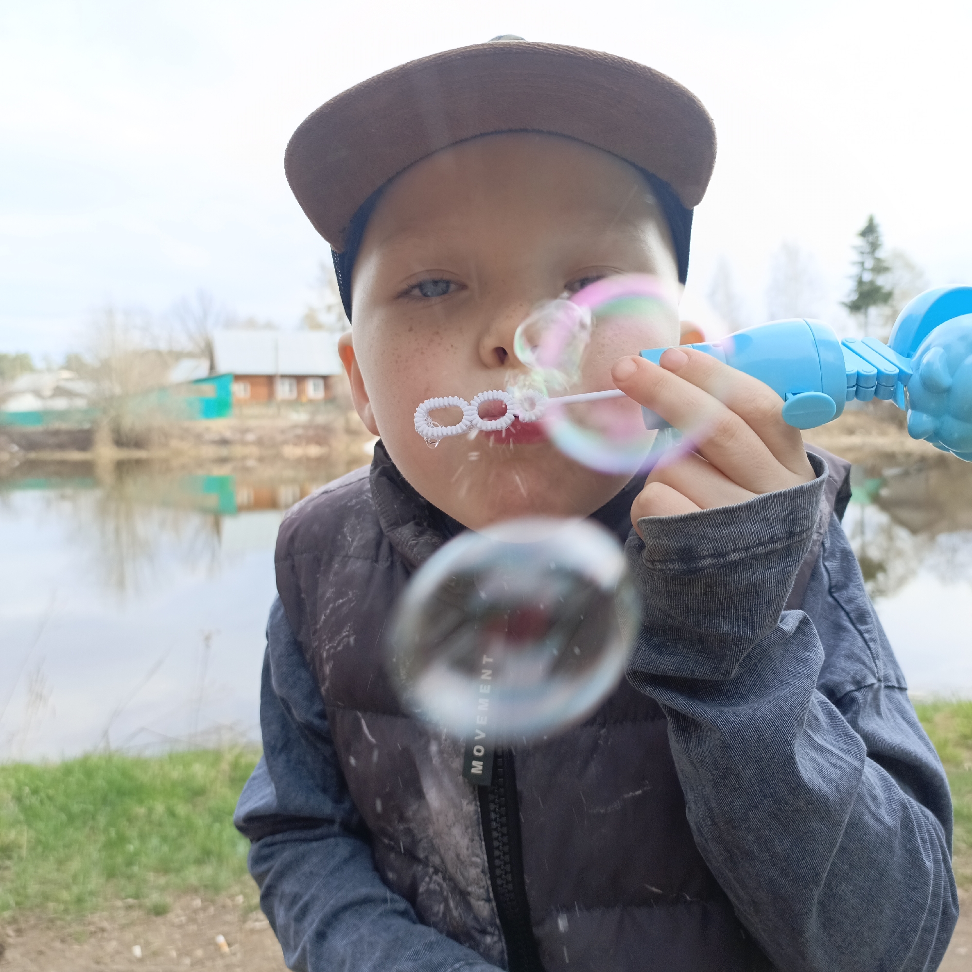 A young boy, appearing to be in elementary school, is featured prominently in a close-up, slightly low-angle shot, actively engaged in blowing bubbles outdoors. He has fair skin, freckles across his nose and cheeks, and light-colored eyes. He wears a brown baseball cap backward, a dark grey or black puffer vest, and a long-sleeved grey denim-like shirt.
He is holding a bright blue bubble gun or blower in his right hand, from which several iridescent bubbles are emerging. One bubble wand, white and circular, is visible near his mouth as if he has just used it to create bubbles. The bubbles are varied in size and reflect a spectrum of colors.
The setting is an outdoor, possibly rural or semi-rural, location beside a calm body of water, likely a river or lake. The water reflects the overcast sky and distant shoreline. Across the water, a few houses with light roofs and some green structures are visible amidst bare trees and patchy vegetation, suggesting a quiet village or hamlet. The immediate foreground shows a grassy bank mixed with some dirt. The sky is uniformly overcast, indicating daytime. The boy's expression is one of concentration and innocent engagement in the playful activity. The word "MOVEMENT" is visible vertically printed on a black tag attached to the zipper pull of his vest.