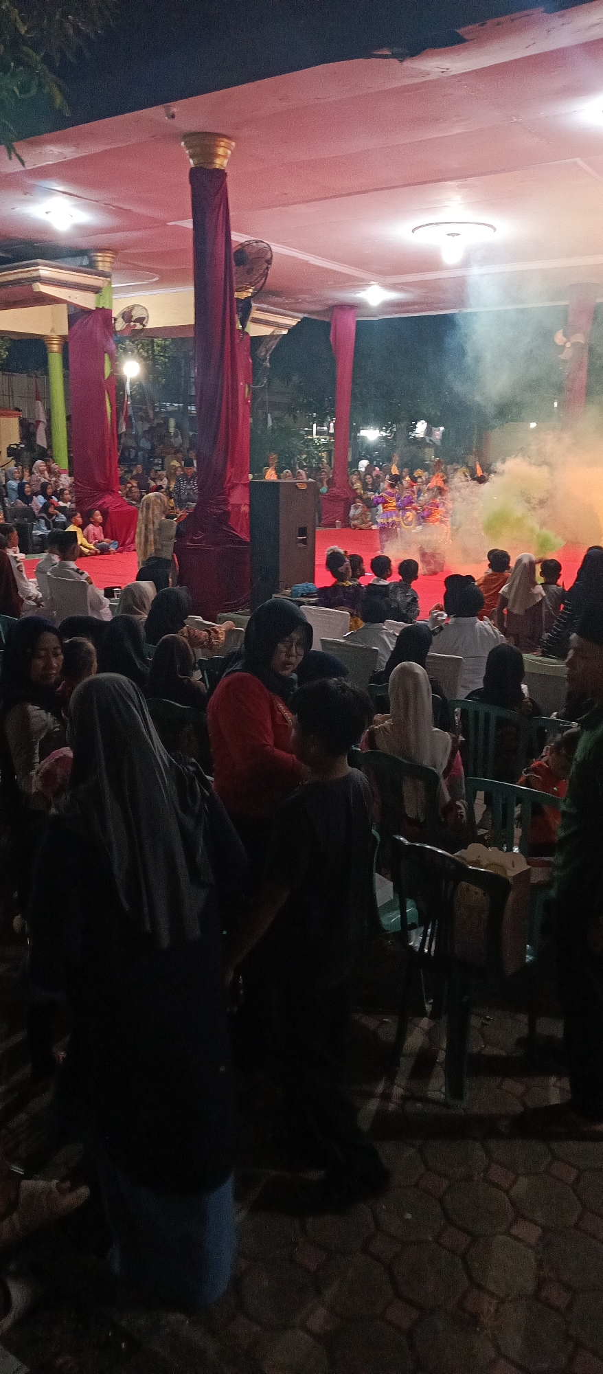 In Arjasa, Indonesia, a stage performance is taking place at night, illuminated by overhead lights and colored smoke effects. A crowd of spectators, many of whom are wearing traditional attire and headscarves, are seated and standing around the performance area. Some are seated on chairs, while others are on the ground. The stage itself is adorned with red drapes and large pillars. Dancers in colorful costumes are visible on stage, surrounded by the smoke. In the foreground, several individuals are captured from the back, observing the performance. The ground is paved with hexagonal stones.