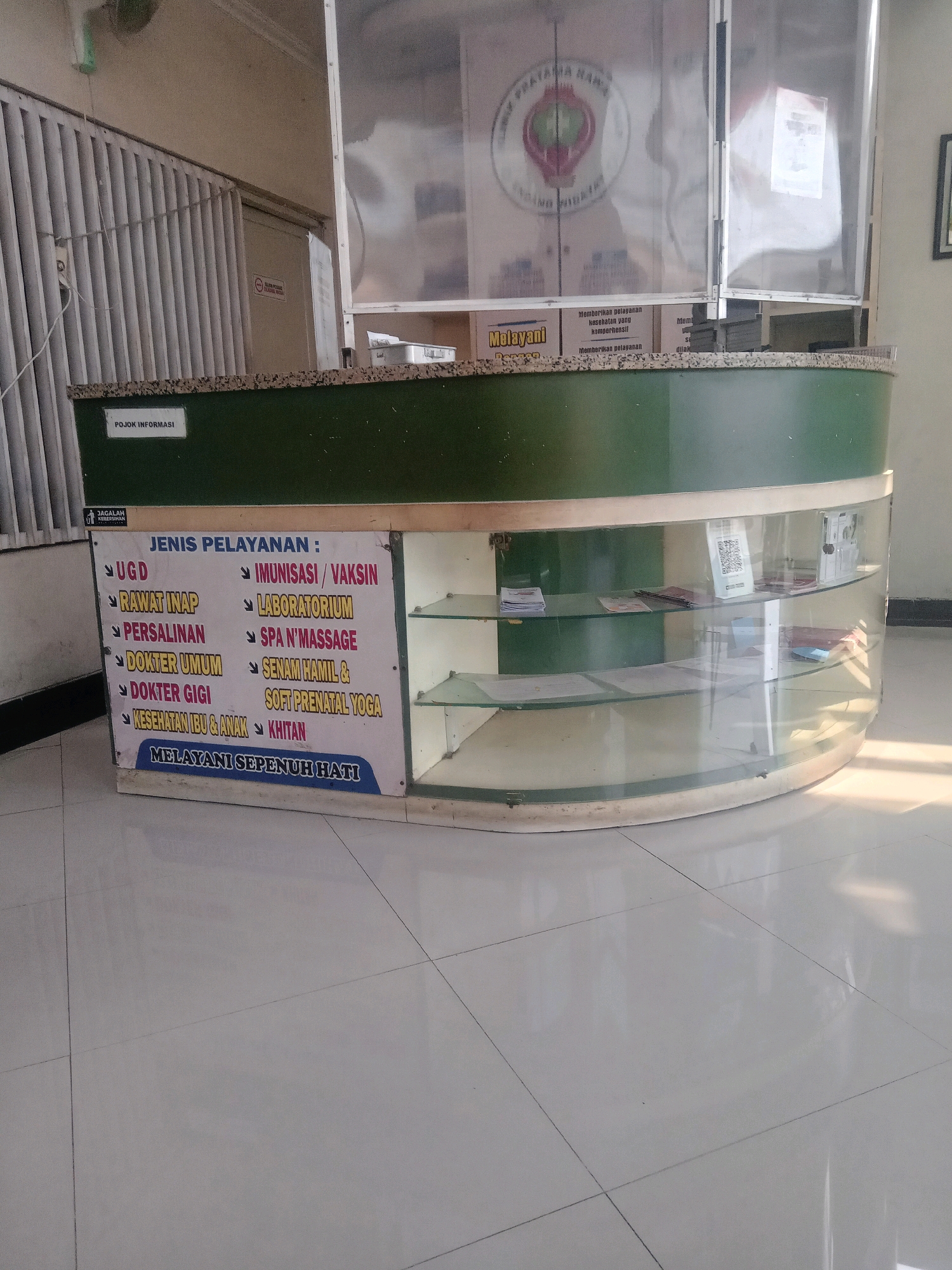 The image shows a reception desk in what appears to be a medical facility, possibly a clinic or hospital, in Pepelegi, Indonesia. The desk is green with a reflective countertop and a glass display area. A sign on the desk lists various services offered, including UGD (Emergency Room), rawat inap (inpatient care), persalinan (childbirth), dokter umum (general practitioner), dokter gigi (dentist), kesehatan ibu & anak (maternal and child health), imunisasi/vaksin (immunization/vaccination), laboratorium (laboratory), spa n' massage, senam hamil & soft prenatal yoga, and khitan (circumcision). The phrase "Melayani Sepenuh Hati" (Serving with All Our Heart) is prominently displayed. Behind the desk, a semi-transparent screen displays a logo and text, likely related to the institution. The floor is tiled and reflects the light. The overall impression is one of an organized and functional service counter within a healthcare setting.