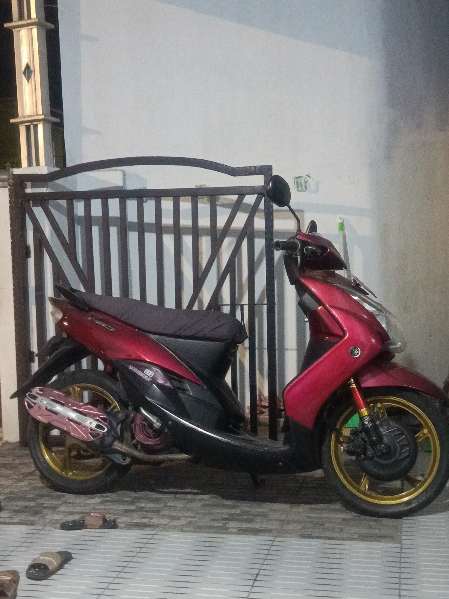 A red scooter is parked on a tiled patio in front of a metal fence. The scooter has a black seat and gold wheels. A pink and silver exhaust pipe is visible. Several pairs of sandals are on the patio. In the background, a white wall and a pillar with lights are visible. The scene is likely in Gorontalo, Indonesia, given the prompt.