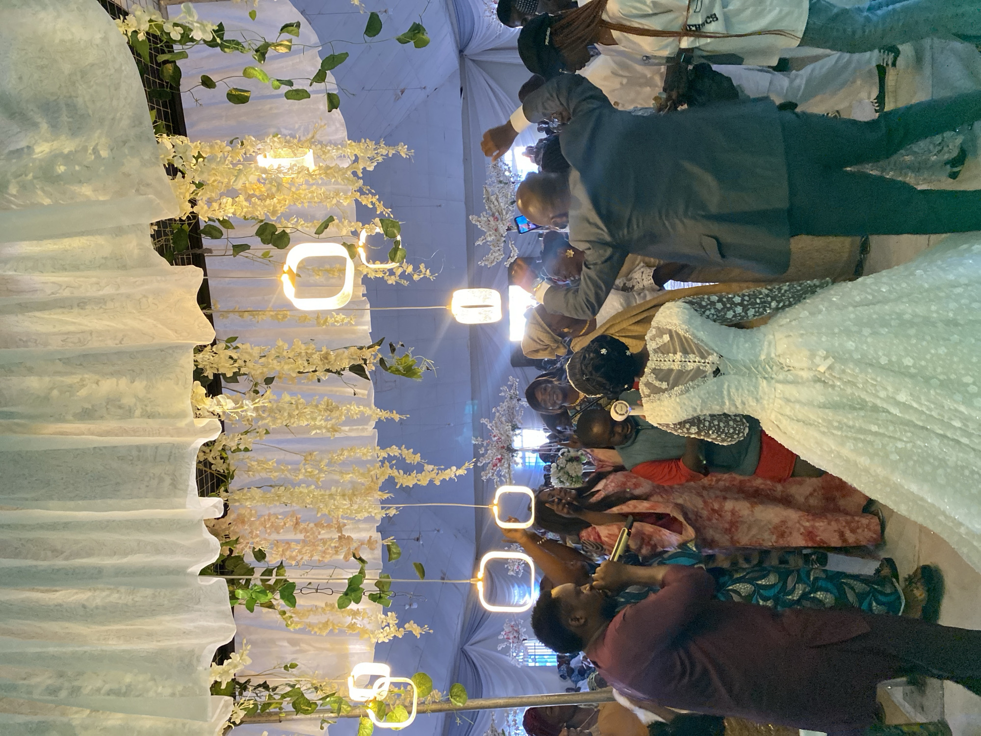 The image captures a scene, likely at an event, in Vom, Nigeria. The foreground features a decorated structure with white fabric draping, adorned with white and yellow floral arrangements, and illuminated by square-shaped light fixtures hanging from above.
Several people are present. A man in a maroon shirt holds a golden object, possibly a microphone, close to his mouth. Others are gathered around, including a man in a suit using a mobile phone to take pictures. A woman in a white dress is partly visible.
The overall atmosphere appears festive. The subjects are engaging in activities related to the event. The environment shows a decorated space, possibly a venue for celebrations.