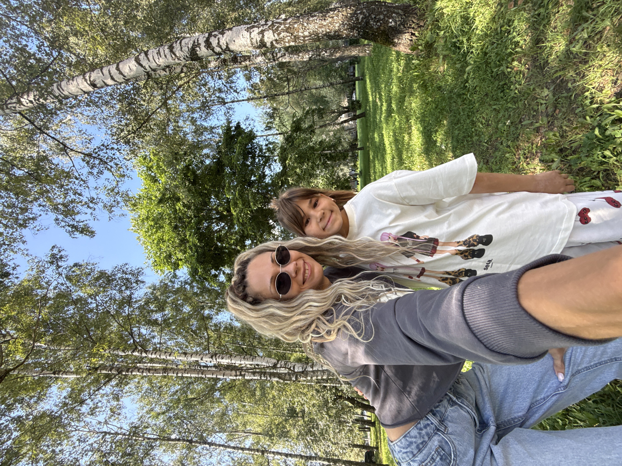 Two females, an adult and a child, are taking a selfie outdoors in a park. The adult, with blonde, curly hair pulled up in a ponytail and wearing sunglasses, is in the foreground, leaning in towards the camera. She is wearing a gray sweatshirt and light blue jeans. The child, with dark hair, is standing to her right, smiling at the camera. She is wearing a white t-shirt with graphics on it. They are surrounded by trees, predominantly birch trees with their distinctive white bark, and lush green grass. The sun is shining, casting dappled light and shadows on the ground. The scene appears to be in Rusanovo, Russia, given the presence of birch trees and the general appearance of the park.