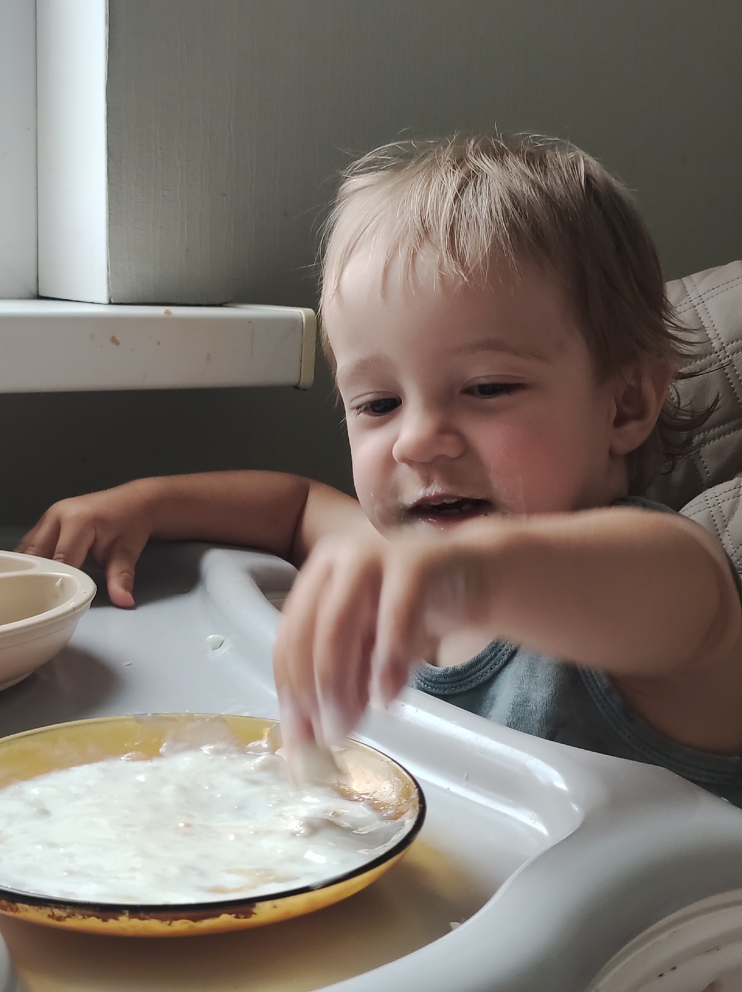 A young child with blonde hair is sitting in a high chair and eating from a yellow bowl filled with a white substance, possibly yogurt or porridge. The child's face is smeared with the food, and they are actively dipping their hand into the bowl. The high chair is white and appears to be made of plastic. In the background, a window sill with a white ledge is visible, along with a textured grey wall. The overall scene suggests a mealtime moment in a home environment, possibly in Chaikovsky, Russia.