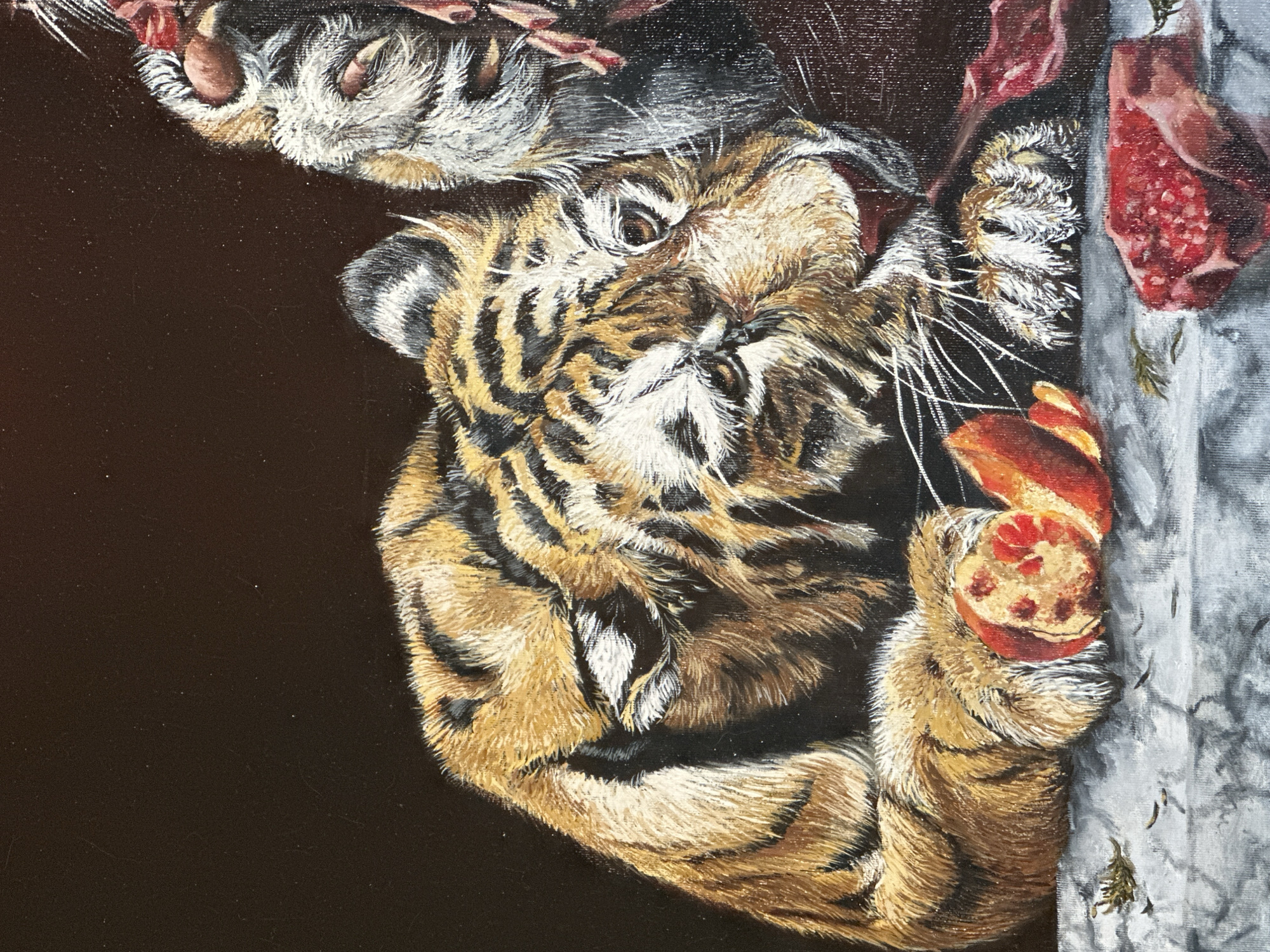 An oil painting on canvas depicts a tiger in close-up, with its head turned to the right, looking down. The tiger's fur is rendered with meticulous detail, showcasing its orange, black, and white stripes. Its paws are resting on a stone-like surface, and it appears to be interacting with pieces of raw meat and a halved pomegranate. The background is a dark, solid color, providing a stark contrast to the vibrant subject. The lighting is dramatic, with highlights and shadows emphasizing the texture of the fur and the materiality of the objects. The overall impression is one of raw natural beauty and predatory focus, presented in a still-life composition.