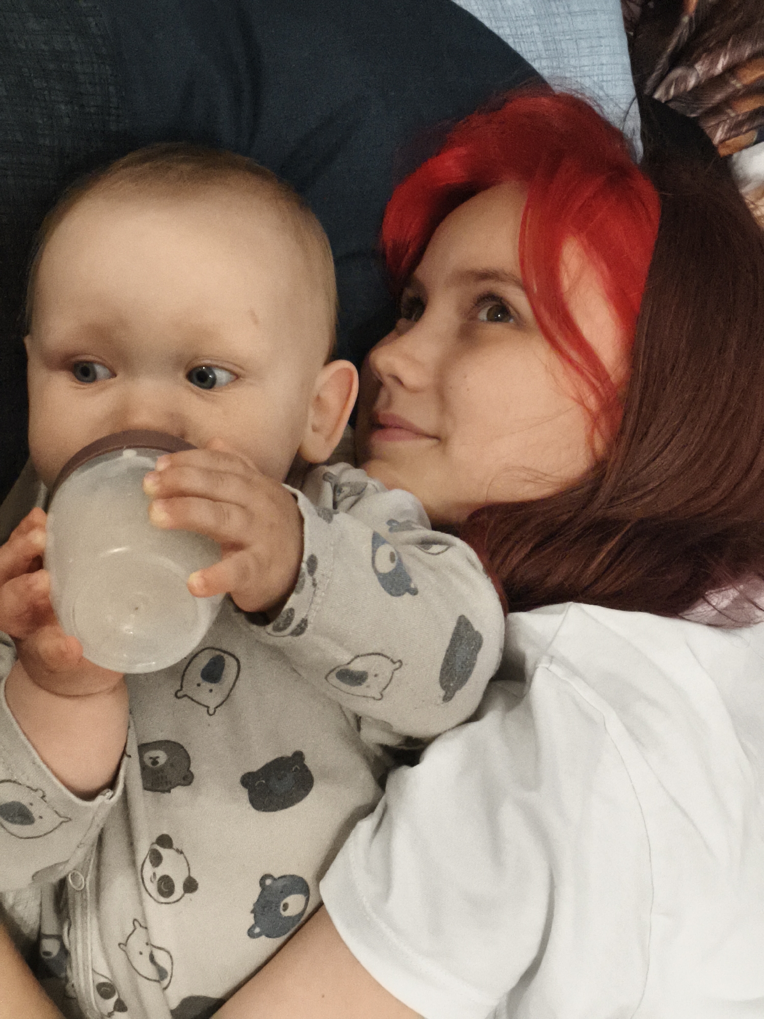 The image shows a close-up of a baby and a young woman lying down. The baby, positioned on the left, is wearing a light gray onesie with a pattern of cartoon bears and pandas. The baby is holding a clear baby bottle filled with a light-colored liquid and drinking from it. The baby has blue eyes and is looking to the left of the frame.
The young woman is on the right, lying next to the baby. She has bright red hair with some darker undertones and is wearing a white t-shirt. Her head is turned towards the baby, and she appears to be looking at the baby with a gentle expression. Her right arm is around the baby.
The background is out of focus but appears to be a bed or sofa, with dark blue and light blue fabric visible. The lighting suggests it is daytime, possibly indoors. The overall mood of the image is intimate and tender, capturing a moment of care and connection between the woman and the baby. There is no visible text in the image. The setting is implied to be a domestic environment in Chaikovsky, Russia.