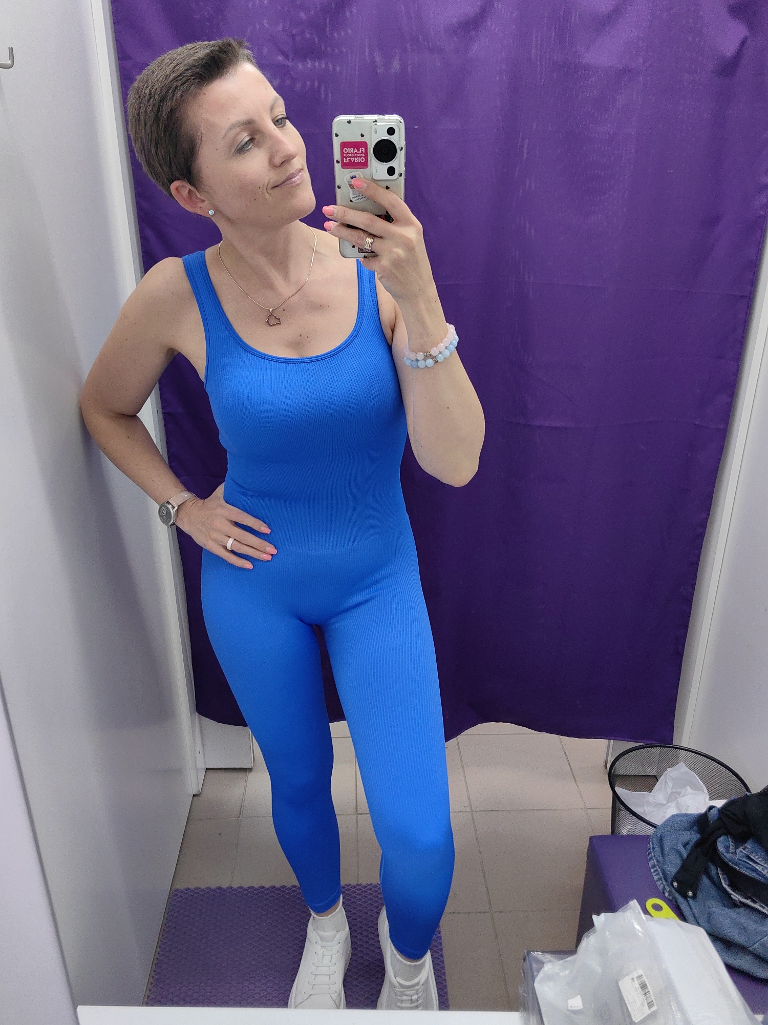 A woman with short brown hair and a pink manicure is taking a selfie in a changing room. She is wearing a vibrant blue ribbed bodysuit and white sneakers. A silver watch is on her left wrist, and she has a thin necklace with a small pendant. She is leaning against a white wall on the left side of the frame, with her right hand on her hip. Behind her, a purple curtain hangs against a light-colored wall. The floor is tiled, and a purple mat is visible under her feet. In the background, a trash can and a purple ottoman with clothing are present. The overall impression is that of a retail fitting room, possibly in Fanipal, Belarus, given the context.