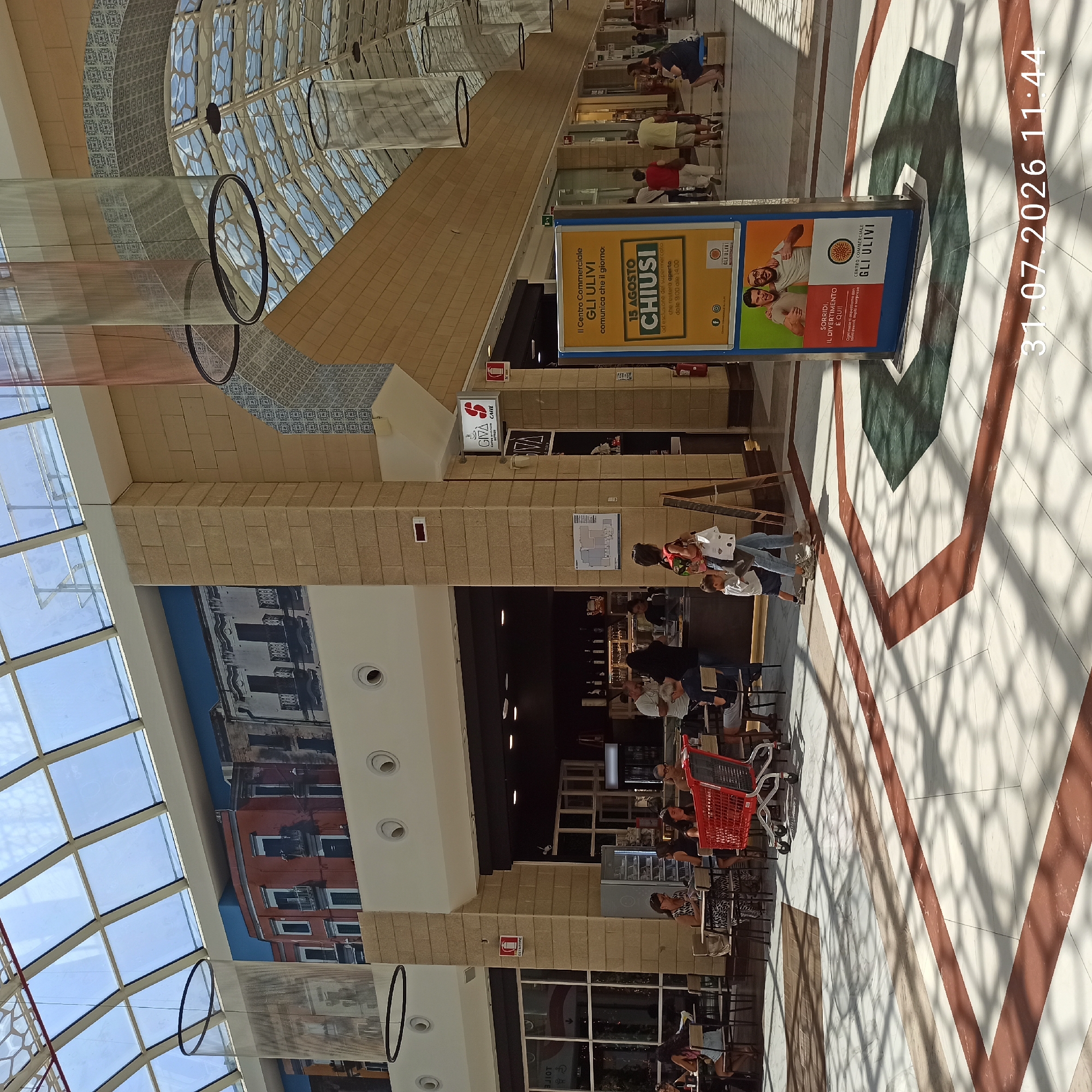 This image shows the interior of the "Gli Ulivi" shopping center in Modugno, Italy. The scene is brightly lit by natural light filtering through a large, segmented glass ceiling. In the foreground, the floor is a patterned marble with geometric designs in red and green. A red shopping cart is positioned prominently.
Several people are visible in the mid-ground, either sitting at tables in an outdoor cafe area or walking through the shopping center. Some are engaged in conversation, while others are carrying bags. The cafe area features several tables and chairs.
A large promotional sign is located on the right side of the image, advertising a special opening day on August 15th. The sign features images of people and text in Italian. Behind the cafe area, store fronts are visible, including one with a sign that reads "GIVA CAFE". The walls of the shopping center are constructed with large, light-colored stone blocks. Decorative elements, including patterned tiles and hanging cylindrical fabric structures, add to the interior design.
The overall impression is of a bustling, modern shopping environment. The timestamp on the bottom right of the image indicates July 31, 2026, at 11:44 AM.