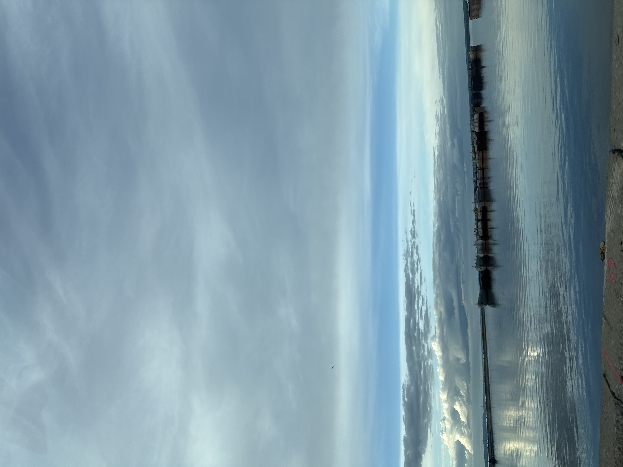 The image depicts a body of water with a harbor or dock area in the distance. The sky is cloudy with varying shades of gray and blue. The water reflects the sky, creating a mirror-like effect. In the distance, there are structures that appear to be part of a port or industrial area. The photograph seems to be taken from a slightly elevated position, possibly a pier or embankment. The location is the city of Chaikovsky, Russia.