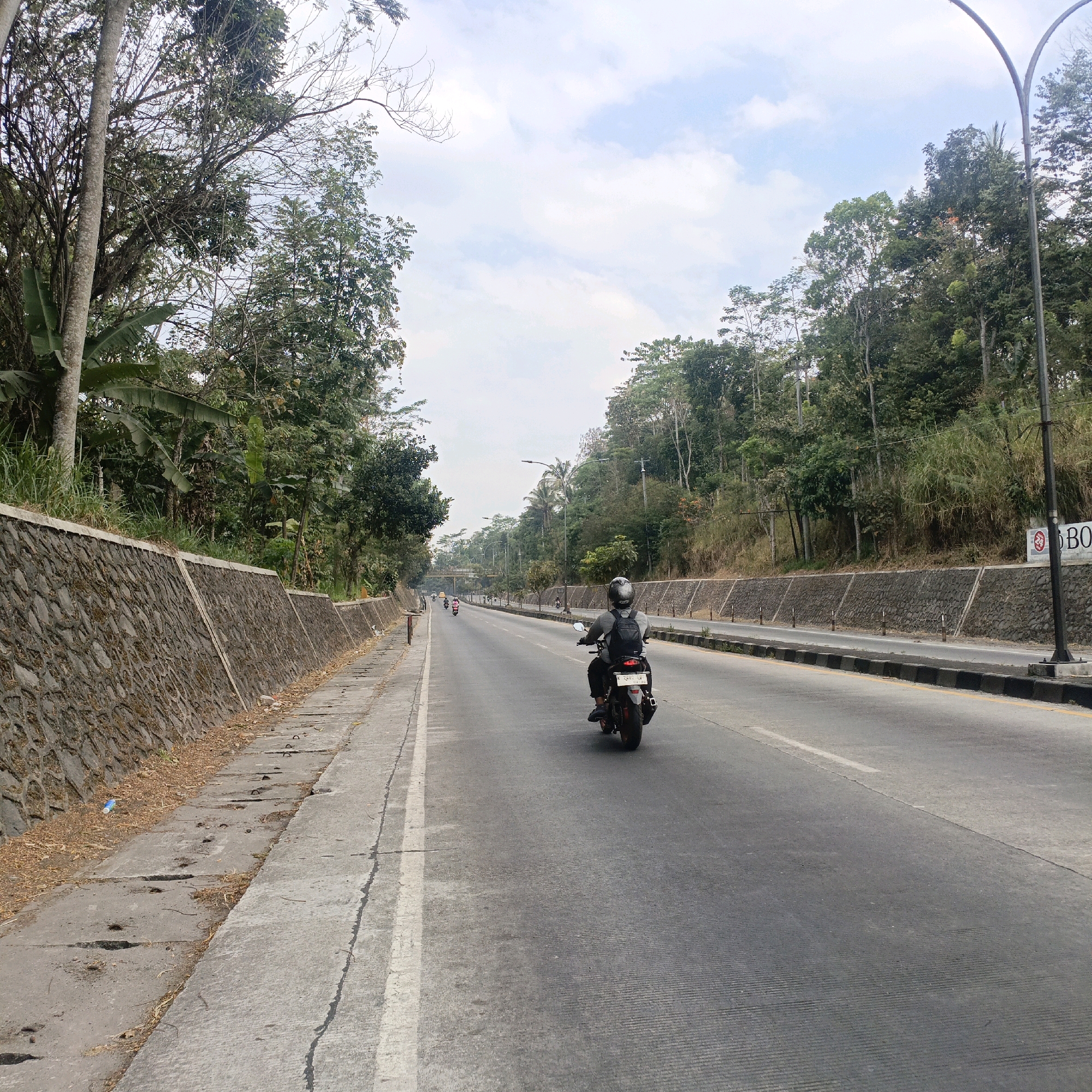 The image shows a two-lane asphalt road with a man riding a motorcycle in the foreground, heading away from the camera. The man is wearing a dark helmet and a dark backpack. The road is flanked by a stone retaining wall on the left and a vegetated hillside on the right. Trees and foliage line both sides of the road, with some distant structures and more trees visible further down the road. The sky is partly cloudy. The scene appears to be a scenic route, possibly in a suburban or rural area, given the natural surroundings and the presence of a paved road. The location is identified as Salatiga, Indonesia, suggesting a tropical or subtropical climate, consistent with the lush vegetation. The road has white lane markings. A street lamp is visible on the right side of the image. In the background, several other motorcycles and a yellow vehicle are also on the road. The retaining wall on the left has a concrete cap and is constructed with irregular stones. The right side of the road has a slightly raised curb and a yellow line.