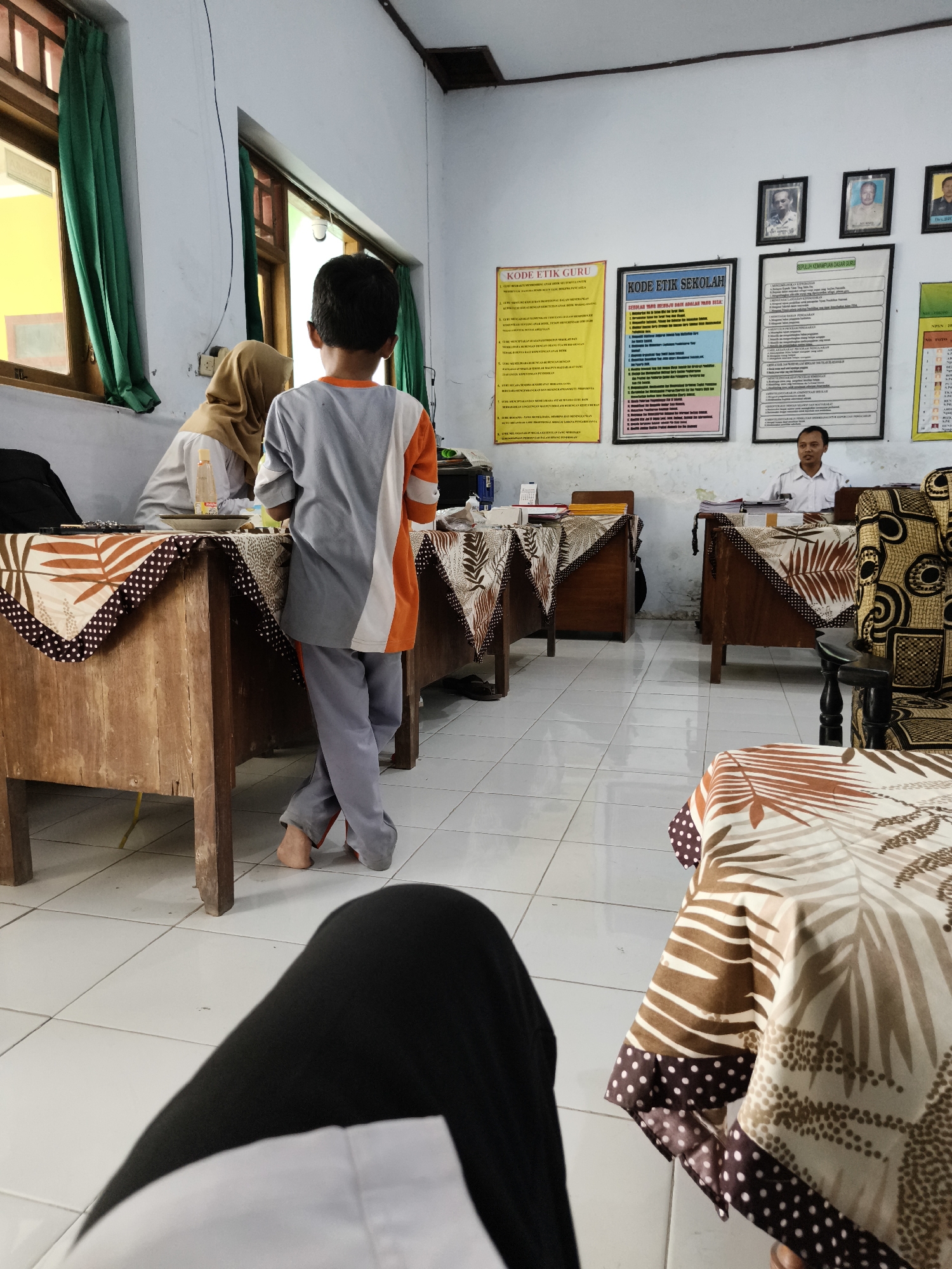 The image depicts a classroom or office space in Betet, Indonesia. In the foreground, a person's legs are visible, suggesting the viewer is seated or has their legs crossed, wearing black pants and a white top.
The room contains several wooden desks, some covered with patterned cloths. On one desk, a woman wearing a hijab is seen interacting with a young boy who is standing facing away from the camera. The boy is wearing a gray, white, and orange shirt and gray pants, with one leg crossed over the other and his bare feet on the tiled floor.
On the walls, several posters and framed pictures are displayed. One poster titled "KODE ETIK GURU" (Teacher's Code of Ethics) and another titled "KODE ETIK SEKOLAH" (School's Code of Ethics) are prominently visible, as are framed photographs of men.
A man in a white shirt is seated behind a desk in the background, appearing to be engaged with some papers. The environment appears to be a functional space, likely associated with an educational institution, given the presence of the code of ethics posters. The floor is tiled, and there are windows with green curtains. The lighting suggests it is daytime, with natural light entering from the windows.
