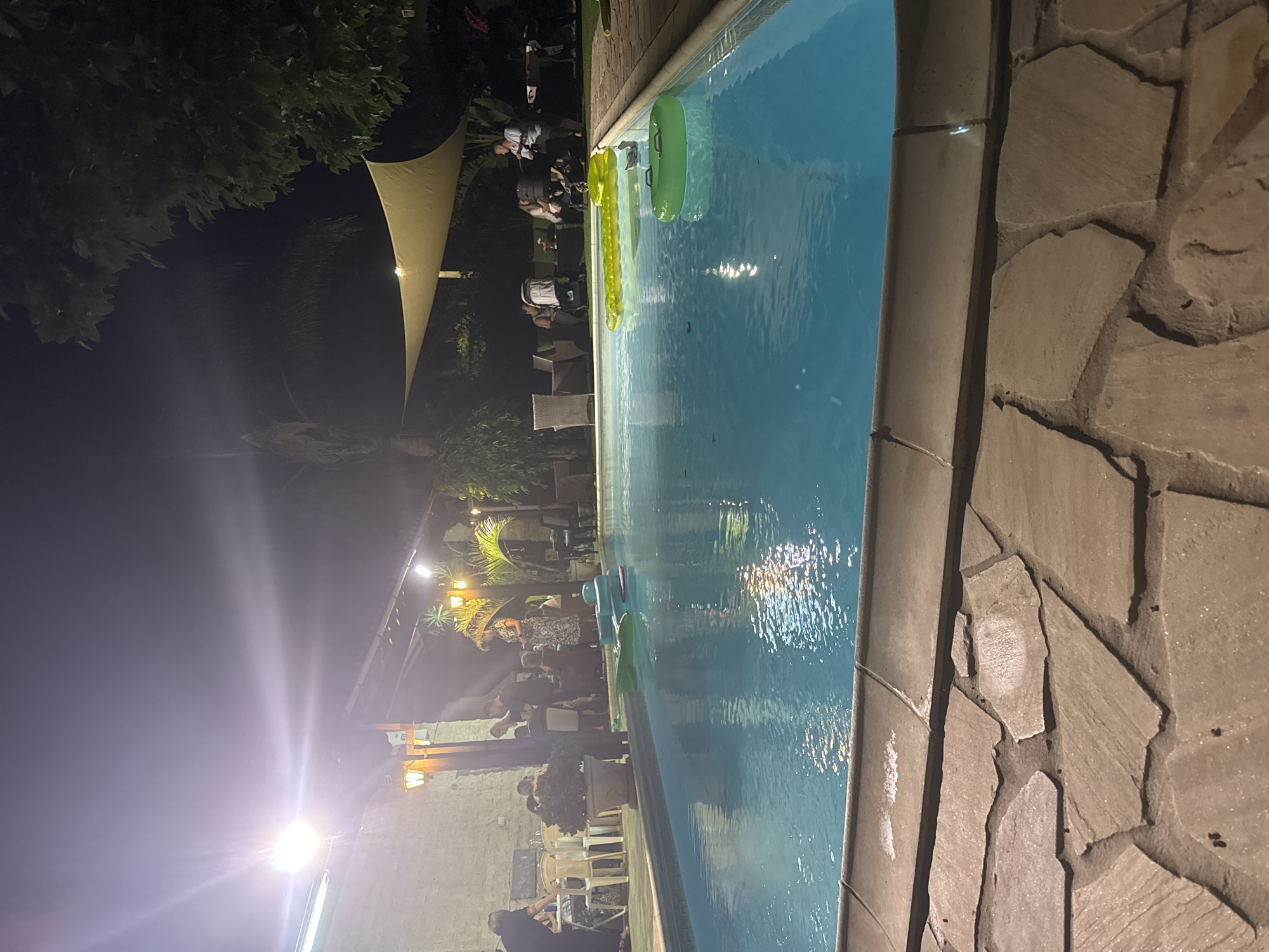 The image shows an outdoor swimming pool at night, likely in Pervolia, Cyprus. The pool is illuminated by artificial lights, creating reflections on the water's surface. Several people are gathered around the pool area, some standing and conversing, while others are seated. There are also inflatable pool toys in the water, including a large yellow raft and a green ring. In the background, there is a covered seating area with tables and chairs, and vegetation, including trees and palm-like plants. Lighting fixtures are visible, casting a warm glow. The overall atmosphere appears to be a social gathering or party.