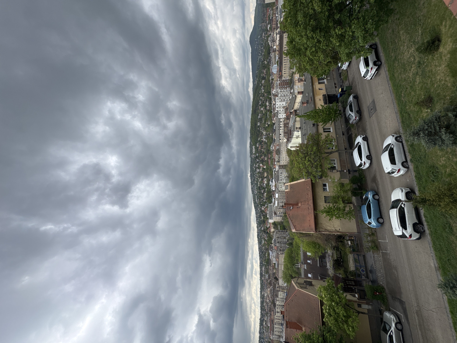 The image presents an elevated panoramic view of a city under a heavily overcast sky. The sky is dominated by thick, textured grey clouds, with some brighter patches suggesting varied light conditions, possibly indicating impending rain or recent stormy weather.
In the midground and background, a dense urban landscape stretches towards distant, hazy hills or mountains. The cityscape is characterized by numerous buildings, including residential houses with visible terracotta roofs, and more substantial multi-story structures further in the distance. Green trees and abundant vegetation are scattered throughout the urban environment, highlighting a lush, possibly spring or summer, setting.
In the foreground and to the right, a paved street or road curves diagonally. Several cars are parked along one side of this street. Most of these vehicles are modern, white SUVs or crossovers. A distinctive light blue car and a silver or light grey vehicle are also visible among them. No people or specific activities are discernible on the street or within the visible parts of the city.
The time of day appears to be daytime, likely late morning or afternoon, judging by the ambient light despite the heavy cloud cover. The scene captures a quiet, somewhat dramatic moment of an urban landscape, consistent with an elevated view over a European city like Budapest, Hungary, on a cloudy day. There is no visible text in the image.