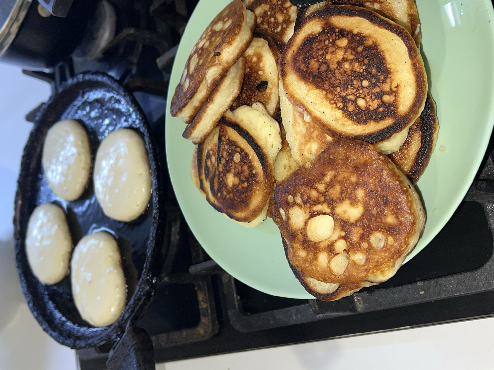 The image captures a culinary scene in what appears to be a kitchen in Noviy, Russia. The main subjects are a collection of freshly cooked, golden-brown pancakes (likely oladyi, a thick Russian pancake) resting on a light green plate, and four portions of uncooked pancake batter still in a cast iron pan on a stovetop.
The activity depicted is the cooking of pancakes. The plate is overflowing with numerous pancakes, displaying various shades of doneness, from lightly browned to deeply caramelized or slightly charred, indicating they were cooked on a hot surface. The texture of the cooked pancakes appears fluffy with visible air pockets.
To the left, a heavy, dark cast iron pan is visible on a black stovetop, likely a gas range given the visible grate structure. Within the pan, four dollops of light, unbaked pancake batter are situated in individual depressions, suggesting a specialized pan for making round, uniform pancakes. The batter is thick and smooth, showing small bubbles. The stovetop surface is dark, contrasting with the white countertop or appliance visible below it. The overall lighting is bright and even, suggesting either ample natural light or good indoor illumination, indicating a daytime setting. There are no people visible, and no text is present in the image.