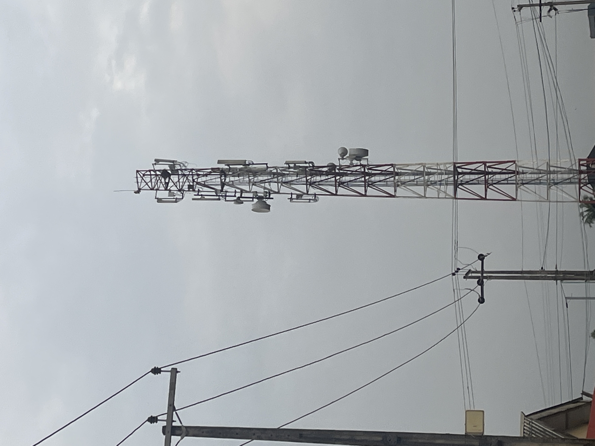 A cell tower, painted in red and white, stands tall against a hazy, overcast sky in Ikorodu, Nigeria. The tower is equipped with multiple antennas and dish-like receivers at various levels. In the foreground, a wooden utility pole with thick black wires stretches across the frame, intersecting with the cell tower. Another smaller utility pole with fewer wires is visible to the right of the cell tower. The lower portion of the image shows the corner of a building with a slanted roof and some faint foliage. The overall scene suggests an urban or semi-urban environment characterized by communication infrastructure and utility lines.