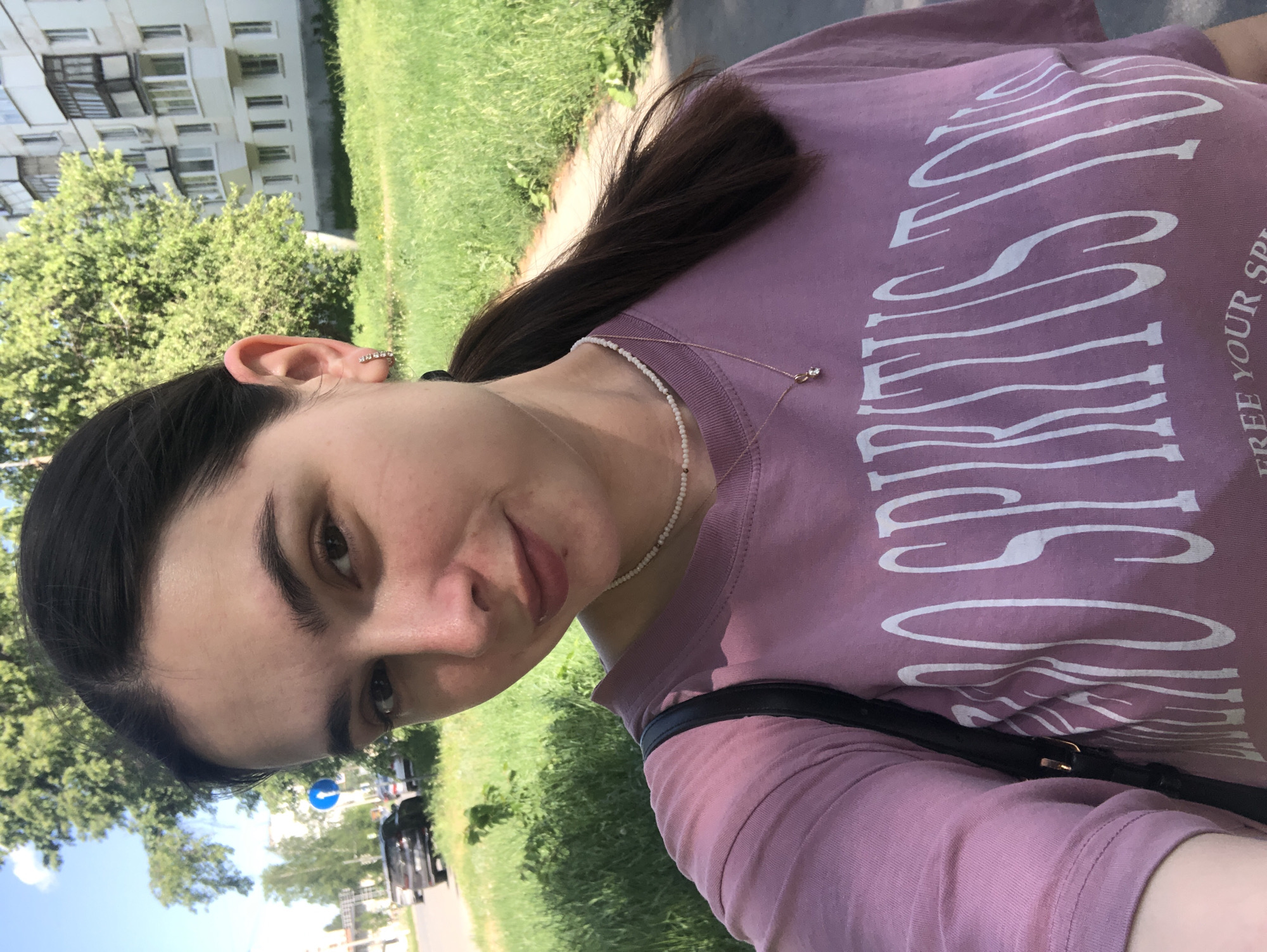 A young woman is centered in the frame, likely taking a selfie, outdoors during the daytime. She has dark hair pulled back, light skin, and is wearing a lilac-colored t-shirt with white lettering. She also has jewelry visible including a beaded necklace and a small gold chain necklace. She is also wearing an earring in her left ear. The background is composed of green grass and trees. In the distance are residential buildings characteristic of urban areas in Russia, specifically the city of Chaikovsky. Cars and street signs are visible, further indicating an urban environment. The overall impression is of a casual, outdoor scene in a Russian city.
