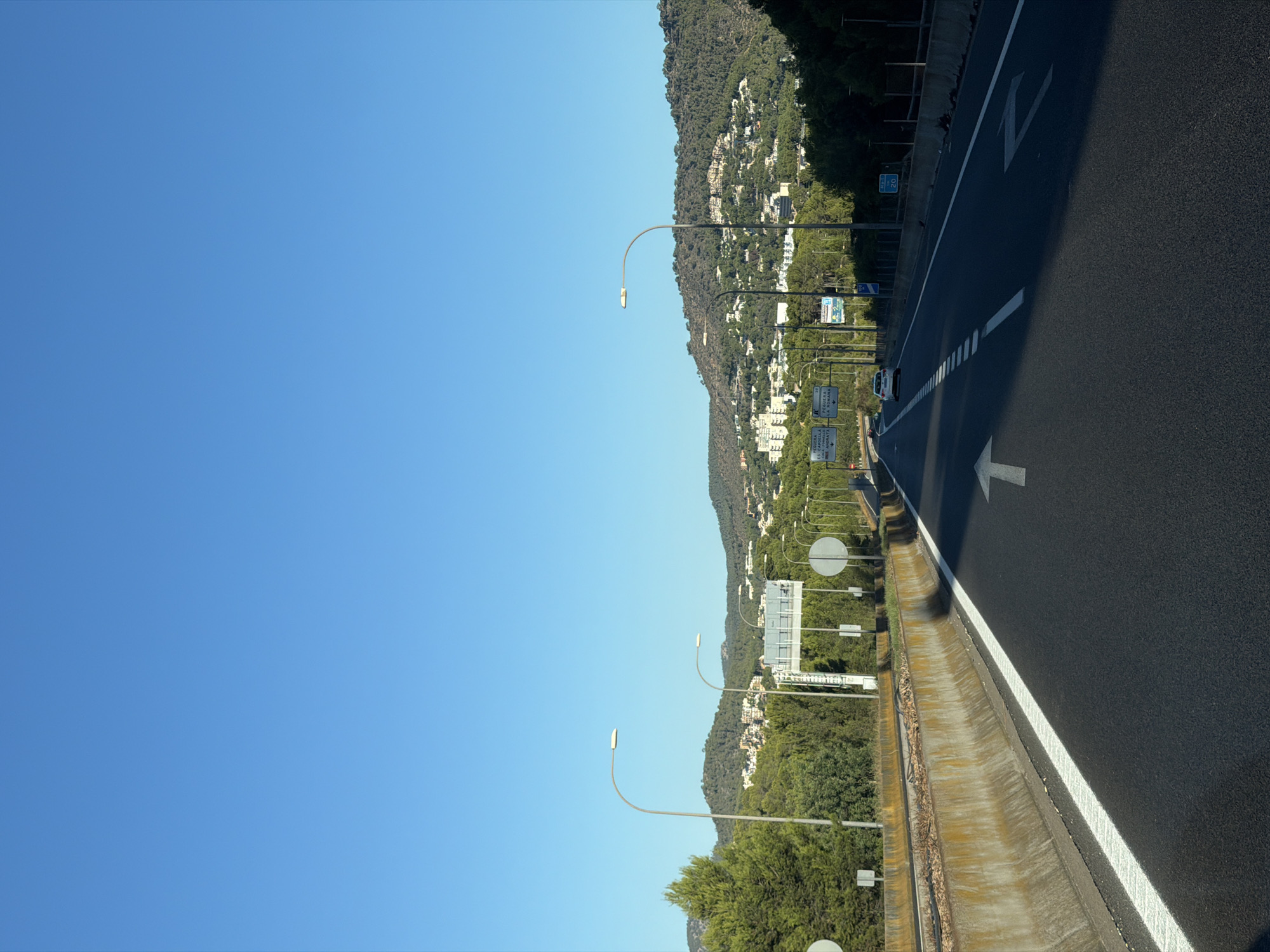 The image shows a view of a highway in the city of Peguera, Spain. The highway is paved and has white lane markings. A concrete barrier separates the highway lanes from an adjacent area with trees and buildings. Several streetlights are visible along the side of the highway. In the background, there are hills covered with trees and buildings, indicating a suburban or semi-rural area. The sky is clear and blue. There is a white car driving on the highway. The scene is a typical highway landscape with a clear blue sky.