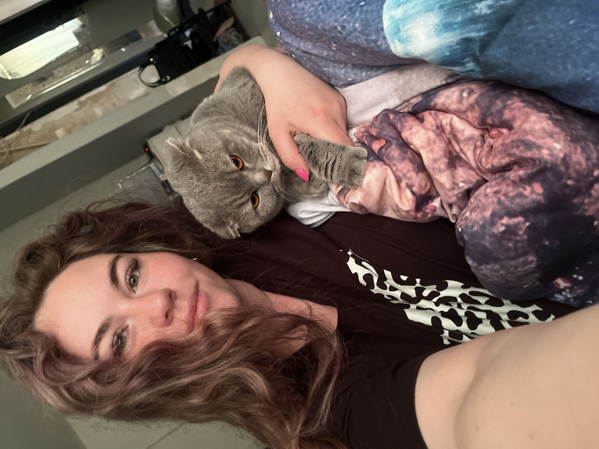 A woman with brown, curly hair is lying down, holding a gray British Shorthair cat. The cat is lying on a colorful blanket. The woman is wearing a dark shirt with a black and white pattern. The background includes a window and various objects. The image appears to be a personal photograph taken indoors, but there is no verifiable evidence to suggest it was taken in the city of Chaikovsky, Russia.