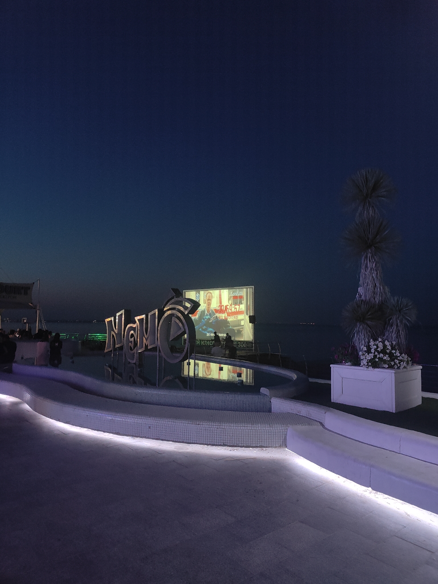 This is a nighttime photograph taken at the "Novi Kino" (New Cinema) outdoor movie theater located on the Odesa embankment in Odesa, Ukraine. The scene depicts the main sign of the cinema, a large, illuminated sculpture in the shape of the words "Novi Kino" with a play button symbol integrated into the "O." The sign is positioned in front of a reflective pool of water. Behind the sign, a large outdoor movie screen displays a scene from a film, featuring a man in a suit and some Ukrainian text. To the right of the screen, a white planter box holds white flowers, and a tall, decorative palm-like tree is visible. In the foreground, there are curved benches and walkways illuminated with subtle purple and white lights. Several people are seated on the benches in the middle ground, facing the screen, suggesting they are attending a movie screening. The background shows the dark expanse of the sea and the city lights in the distance. The sky is a deep, clear blue, indicating it is twilight or early night.