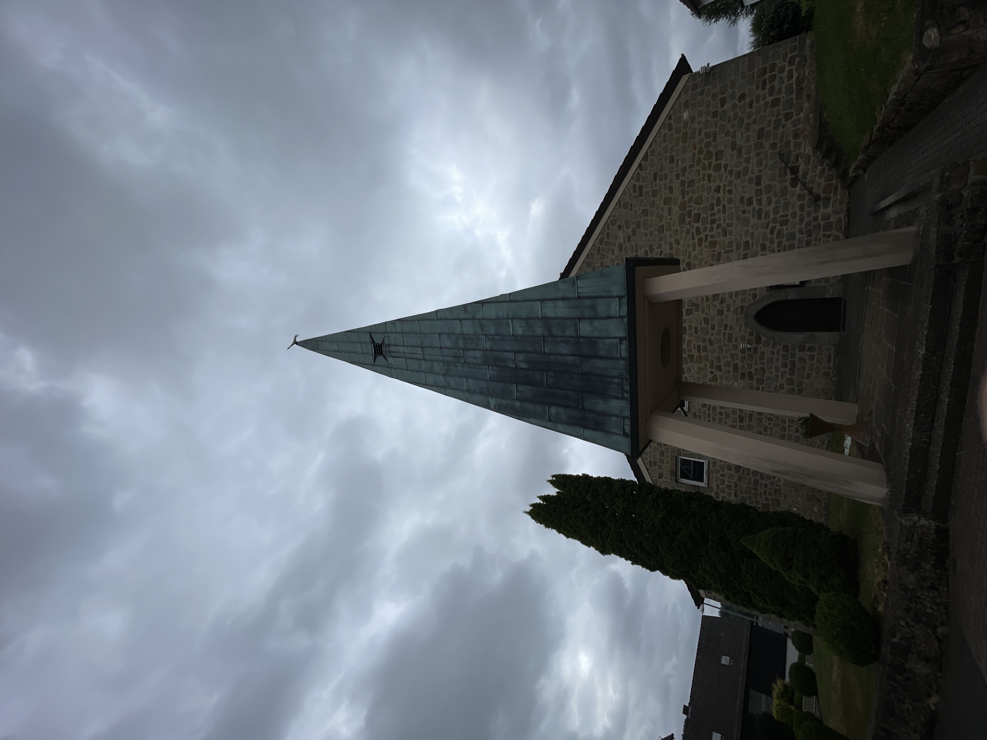 This image depicts a church in Schorbach, Germany. The church features a stone facade and a tall, pointed spire covered in a weathered, greenish-blue material. The spire is supported by angled concrete pillars that extend from the ground to the base of the spire's overhang. A dark, arched doorway is visible on the stone wall beneath the spire. Adjacent to the church, on the left, is a tall, slender evergreen tree. In the background, residential buildings with tiled roofs can be seen. The sky is overcast with heavy, grey clouds. The foreground shows a paved walkway leading to the church entrance, flanked by steps and stone walls. The overall scene suggests a cloudy or impending stormy day.