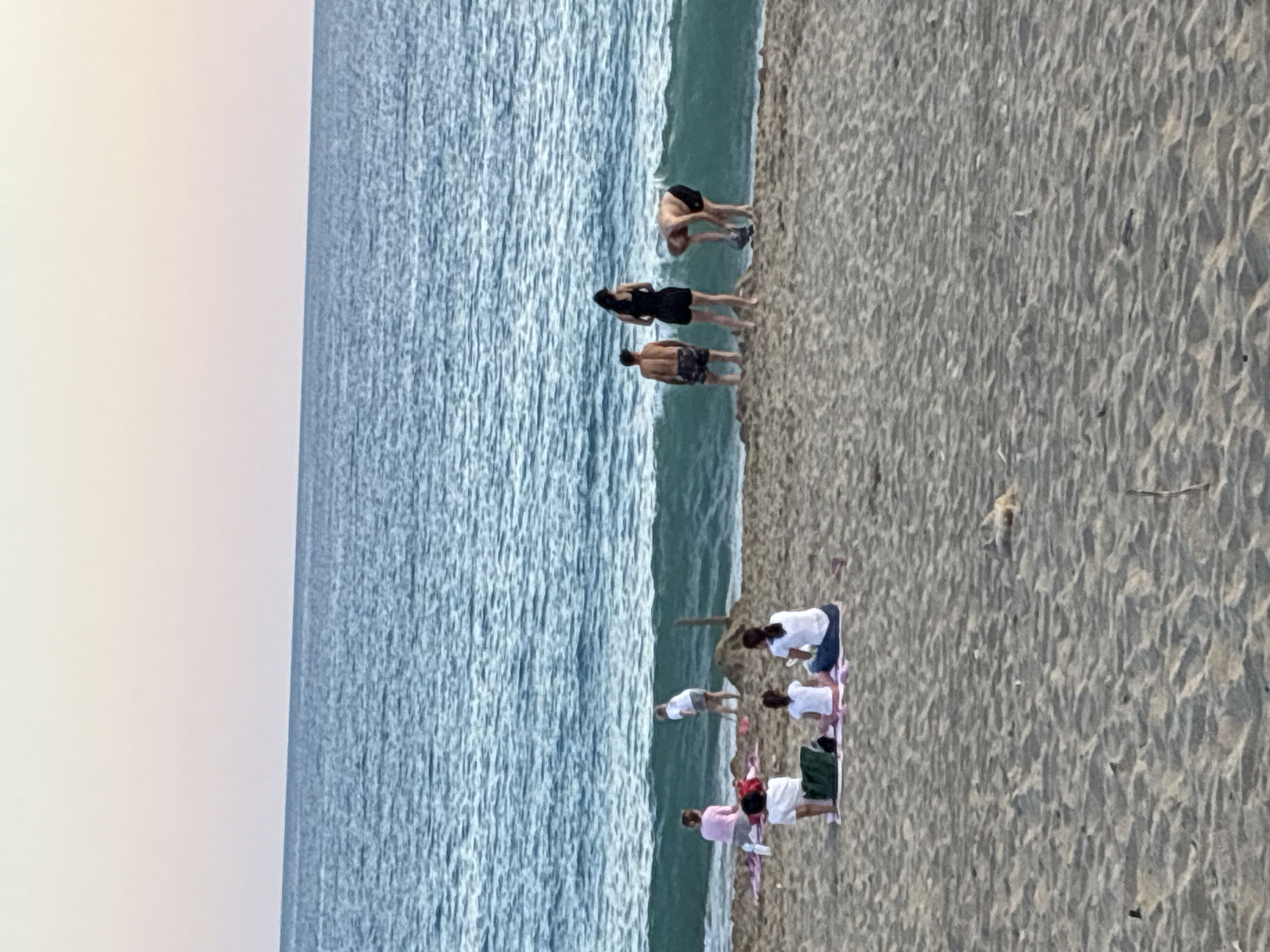 In Alanya, Turkey, on a sandy beach, a group of people are gathered by the sea. Several individuals are sitting on blankets, facing the ocean. In the background, a child is playing with sand and a shovel. Further to the right, three adults are near the water, with one person bending down to pick up something. The ocean is calm with small waves, and the sky is a soft gradient of pink and blue, indicating either sunrise or sunset.