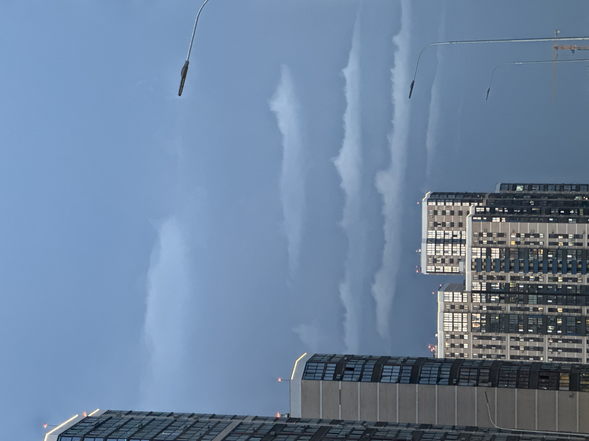 The image shows a view in the city of Krasnodar, Russia. Tall buildings dominate the lower part of the frame, with their windows and architectural details visible. Above the buildings, the sky is overcast with elongated clouds. Two streetlights are positioned on either side of the frame. A construction crane can be seen in the background. The scene appears to be taken at dusk.
