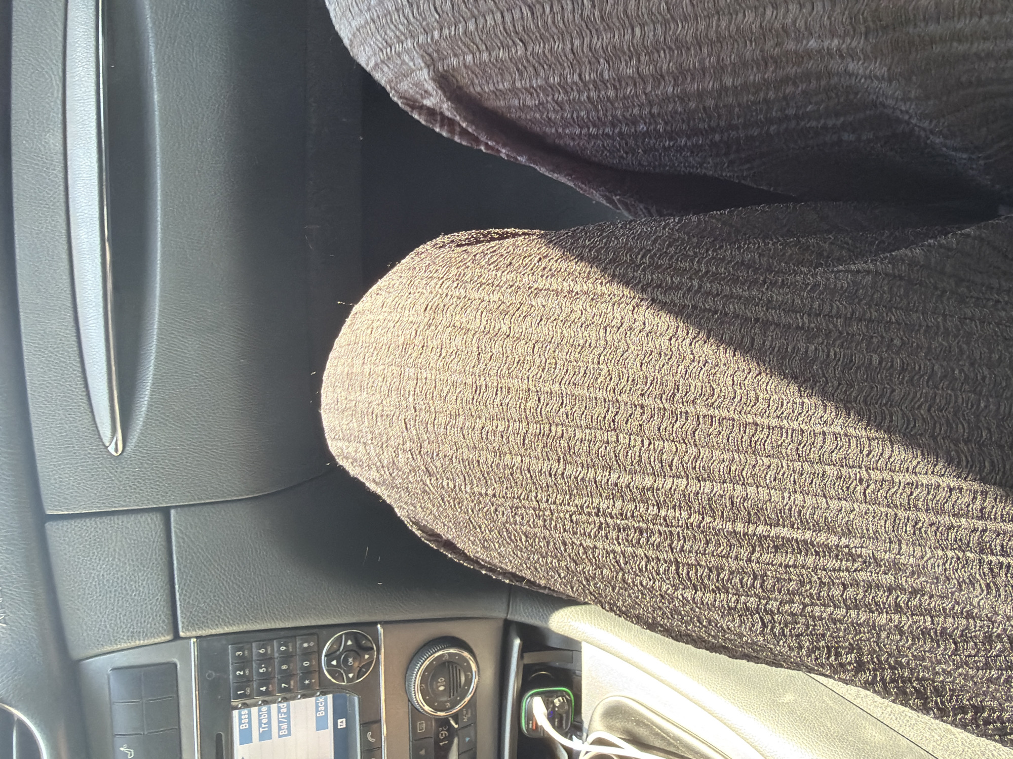 The image shows a close-up view of a person's legs and lap inside a car. The person is wearing dark, textured trousers. The car's dashboard is visible in the background, including the radio and air vents. A phone charger is plugged into a power outlet. The image appears to be taken during the daytime, with sunlight casting shadows on the person's legs. The location is not specified, and there is no indication that it is in Plovdiv, Bulgaria.