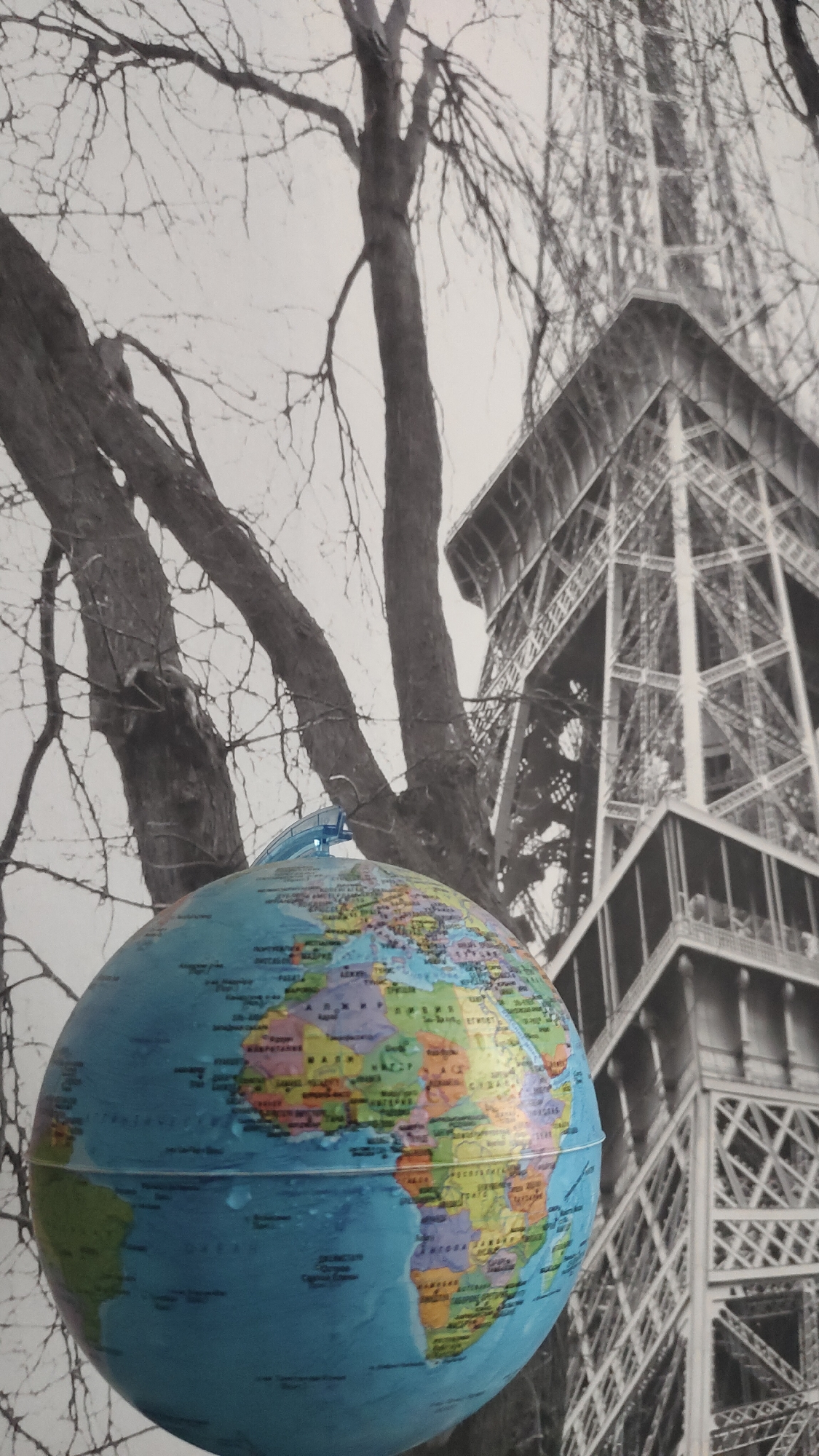 The image shows a globe in front of a black and white background depicting a bare tree and the Eiffel Tower. The globe is predominantly blue, representing the oceans, with colorful continents visible. It's unclear if the scene is a photograph or a composite of different images. There is no indication that this is located in the city of Krasnoyarsk, Russia.