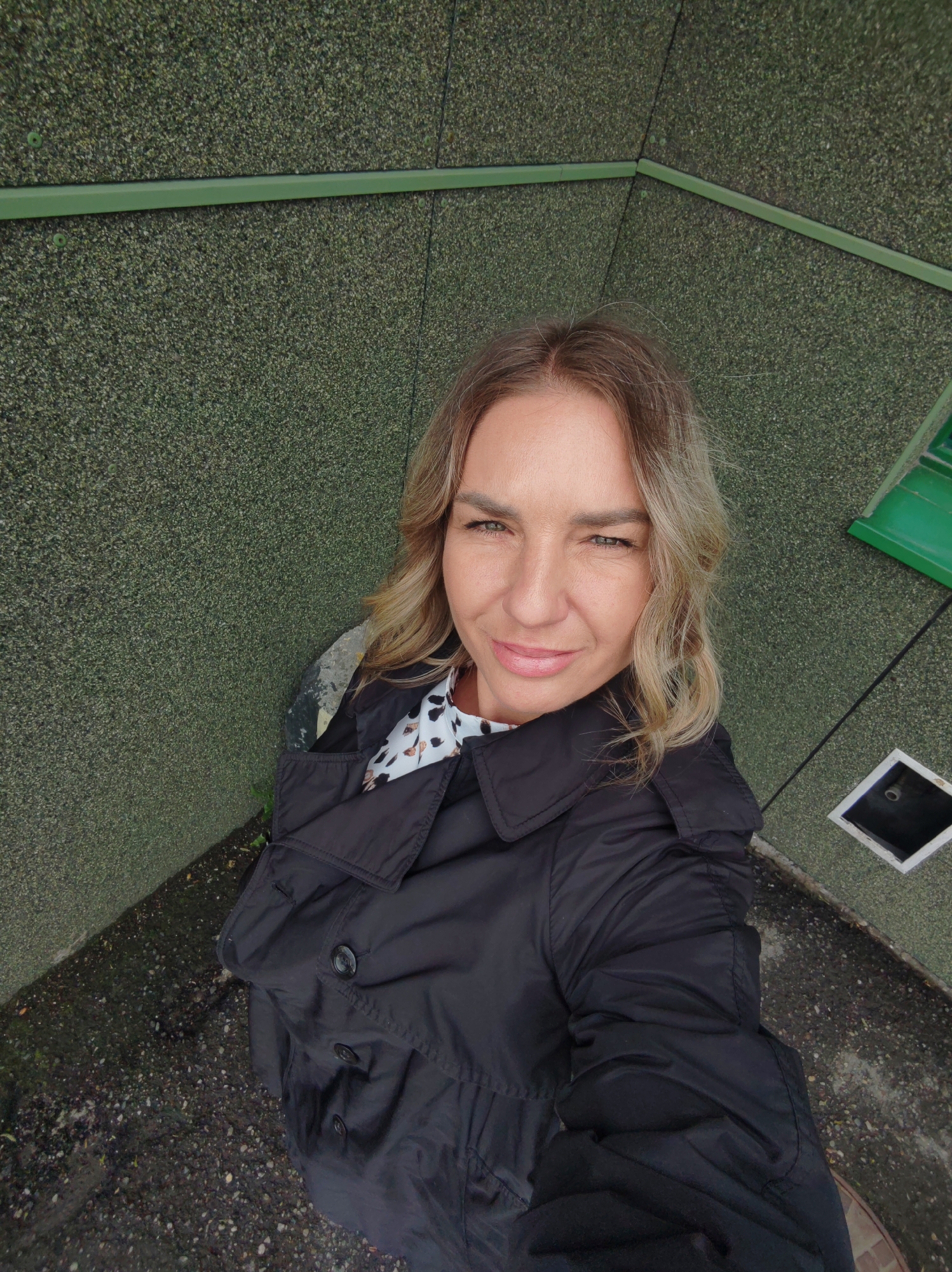 A woman stands in what appears to be a corner of an exterior structure. The wall behind her is textured green with horizontal green metal trim. A green downspout and a square drain are visible to her right. She is wearing a black trench coat over a patterned blouse. The ground is a dark, speckled concrete or asphalt. The image does not provide enough detail to identify the specific location in Petropavlovsk-Kamchatsky.