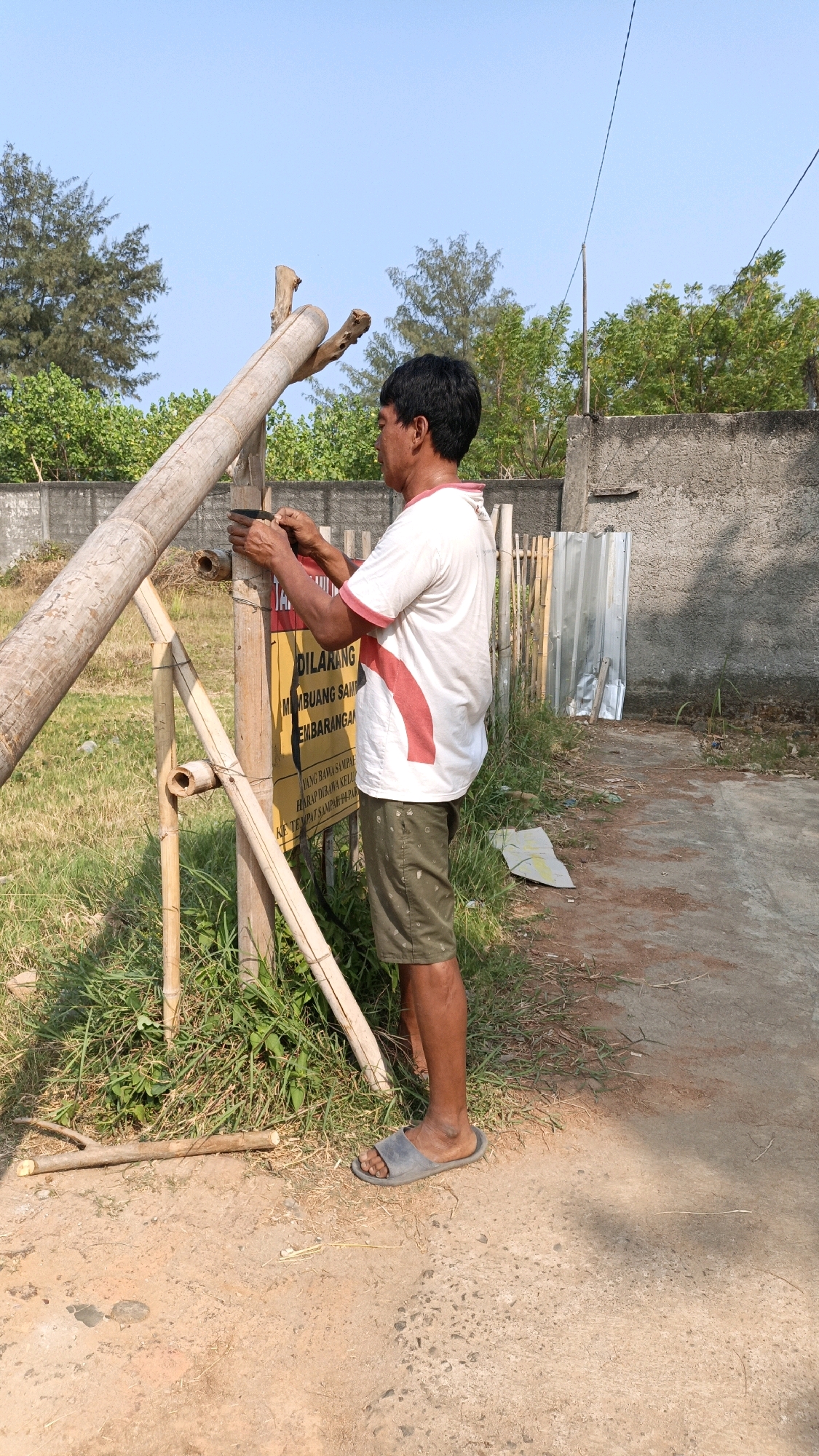 An Indonesian man, standing on a dirt path in Cikal, Indonesia, is in the process of attaching a yellow sign to a makeshift bamboo structure. The man wears a white t-shirt with red accents and olive green shorts, along with gray flip-flops. The sign, predominantly yellow with red and black text, appears to be a warning or informational notice. The surrounding environment is characterized by dry grass, scattered debris, and a concrete wall in the background, indicative of an outdoor, possibly semi-rural or undeveloped area. The sky is clear and blue. The bamboo structure seems to be a gate or entrance marker, with large bamboo poles supporting a smaller vertical piece to which the sign is being affixed.