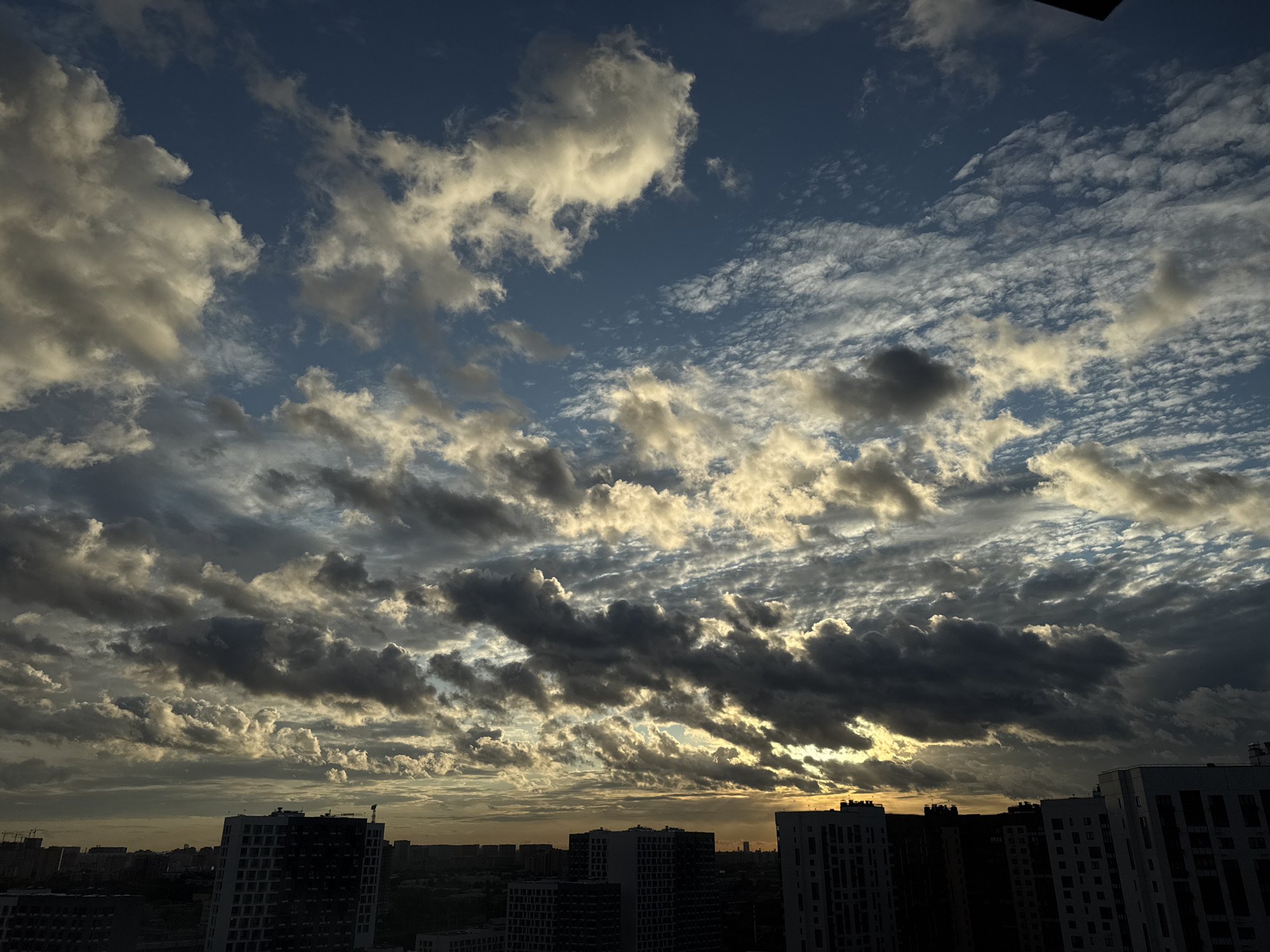 The image depicts a dramatic sky filled with a variety of clouds over the cityscape of Lyubertsy, Russia. The sky is a deep blue, with large, fluffy white clouds interspersed with darker, more ominous formations. Wispy, cirrus-like clouds are also visible, creating a textured effect. Sunlight breaks through in places, illuminating the edges of some clouds with a warm, golden hue, while others are cast in shadow.
Below the sky, the silhouette of a city can be seen. Several modern, multi-story buildings with dark facades and numerous windows dominate the lower portion of the image. Some buildings appear to be residential complexes, with their architecture suggesting they are relatively new constructions. The urban landscape extends to the horizon, where hints of other structures and possibly distant trees are visible against the dusky sky. The overall scene captures a moment of changing weather, possibly during sunrise or sunset, with the sky serving as the primary focal point.