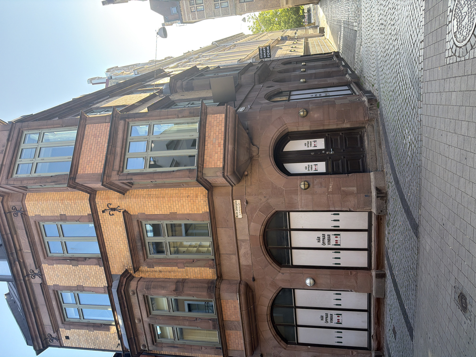 The image shows a street corner in Malmö, Sweden, on a sunny day. The main subject is a corner building with a facade of orange brick and rust-colored stone. The building has several windows with green frames, some of which are bay windows. On the ground floor, there are large arched windows with white posters that say "HÄR ÖPPNAR VINBAR" (Here opens wine bar) and feature illustrations of wine bottles and glasses. A dark wooden door is also visible on the ground floor.
A street sign on the building reads "Mäster Johansgatan" with a smaller sign below indicating a street number. To the right of the building, another sign reads "STUDIO SMART FORM". The street is paved with cobblestones and some modern paving stones. In the background, other buildings and a glimpse of a tree can be seen. The sky is clear and blue. There are no people or vehicles visible in the image.