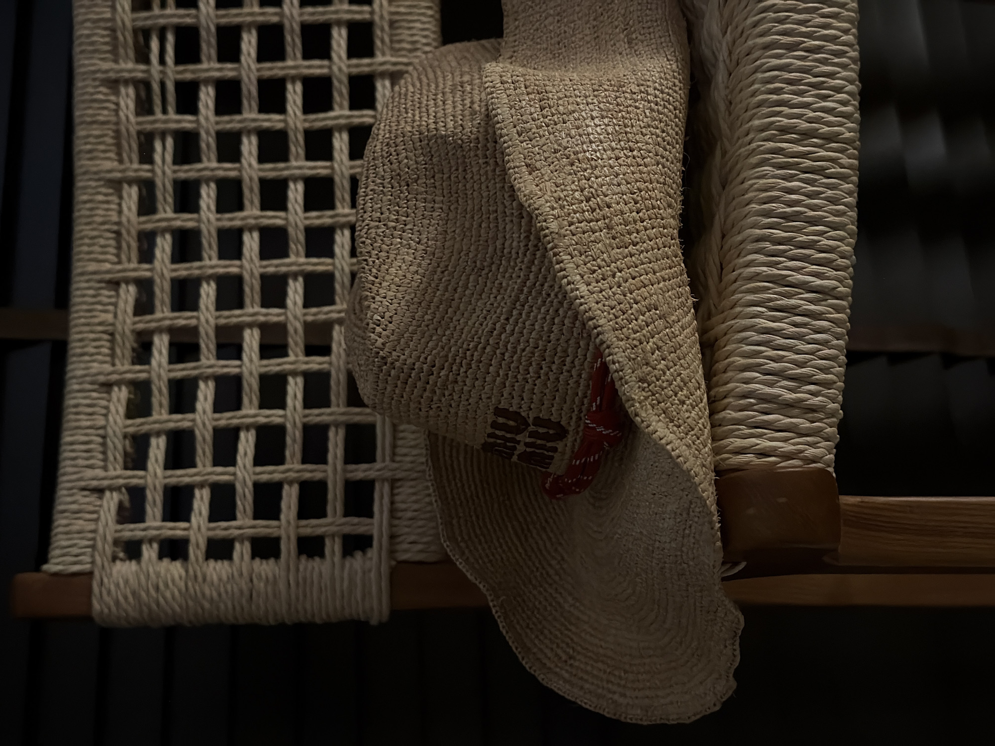 An outdoor photograph taken at Baan Plai Laem, Thailand shows a close-up of a woven straw hat with a red and white patterned band, placed on a woven chair. The hat is angled, with its brim casting a shadow. The chair is made of natural-colored rope woven into a grid pattern and has wooden legs. The background is dark, suggesting it might be evening or a shaded area. The focus is sharp on the hat and the texture of the chair.