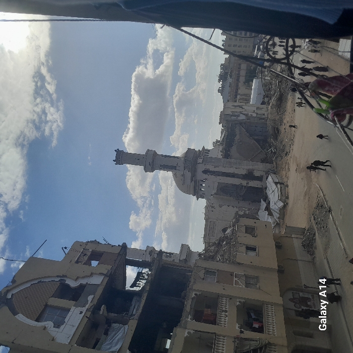 Amidst a blue sky dotted with white clouds, a scene of destruction unfolds. Buildings are heavily damaged, reduced to ruins. A prominent mosque with a tall minaret stands amidst the wreckage. A road stretches alongside the devastation, where people walk or stand. The overall impression is one of devastation and loss.