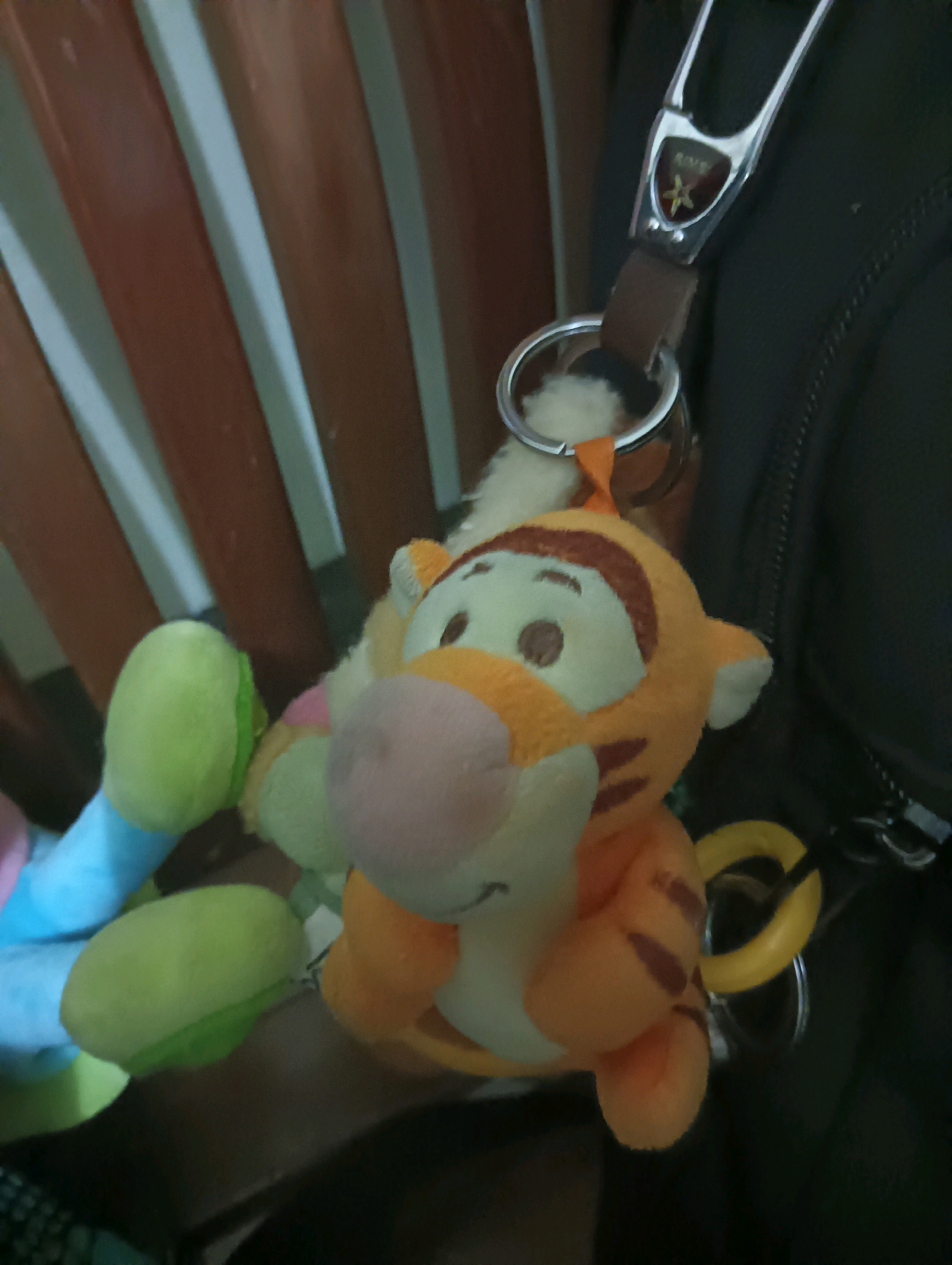 The image shows a close-up view of a Tigger keychain dangling from a dark bag, positioned in front of a wooden chair back. The Tigger plush toy is orange with black stripes and has a pink nose. It is attached to a silver keychain with a brown leather strap and a decorative emblem. Part of another plush toy, possibly a snake, is visible on the left, with sections colored blue and green. The background consists of the wooden slats of a chair and the dark fabric of the bag. The lighting suggests an indoor setting. There are no people or animals present in the immediate vicinity of the objects. The image does not contain any information that would allow for the determination of its location in Tomohon, Indonesia.