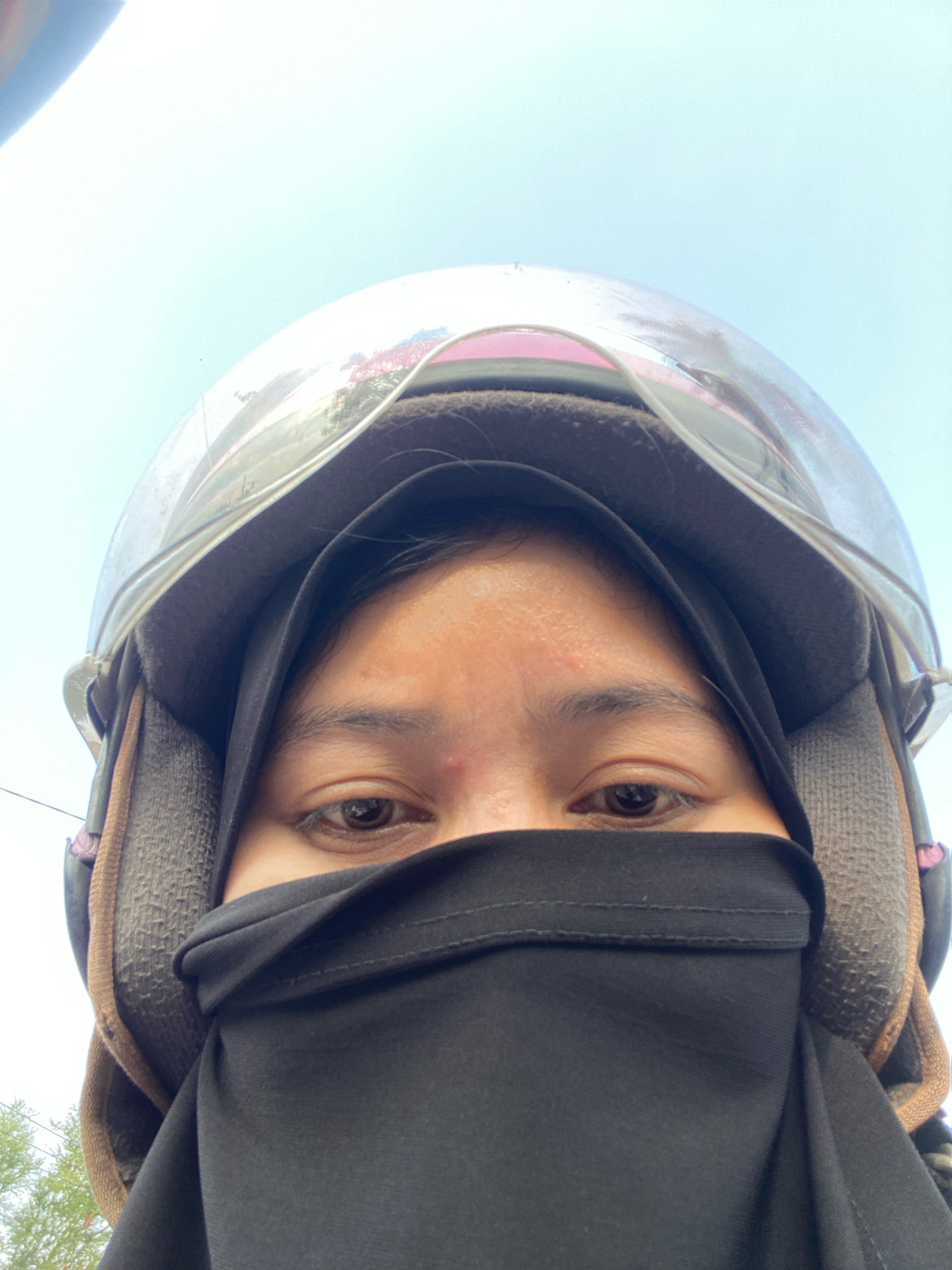 A low-angle, close-up shot shows a person wearing a helmet and a black face covering. The person's eyes are visible, looking directly at the camera. The helmet has a clear visor, reflecting the sky and some greenery. The sky is light blue and overcast. The lower part of the image shows trees. This photo was taken in Banjarbaru, Indonesia.