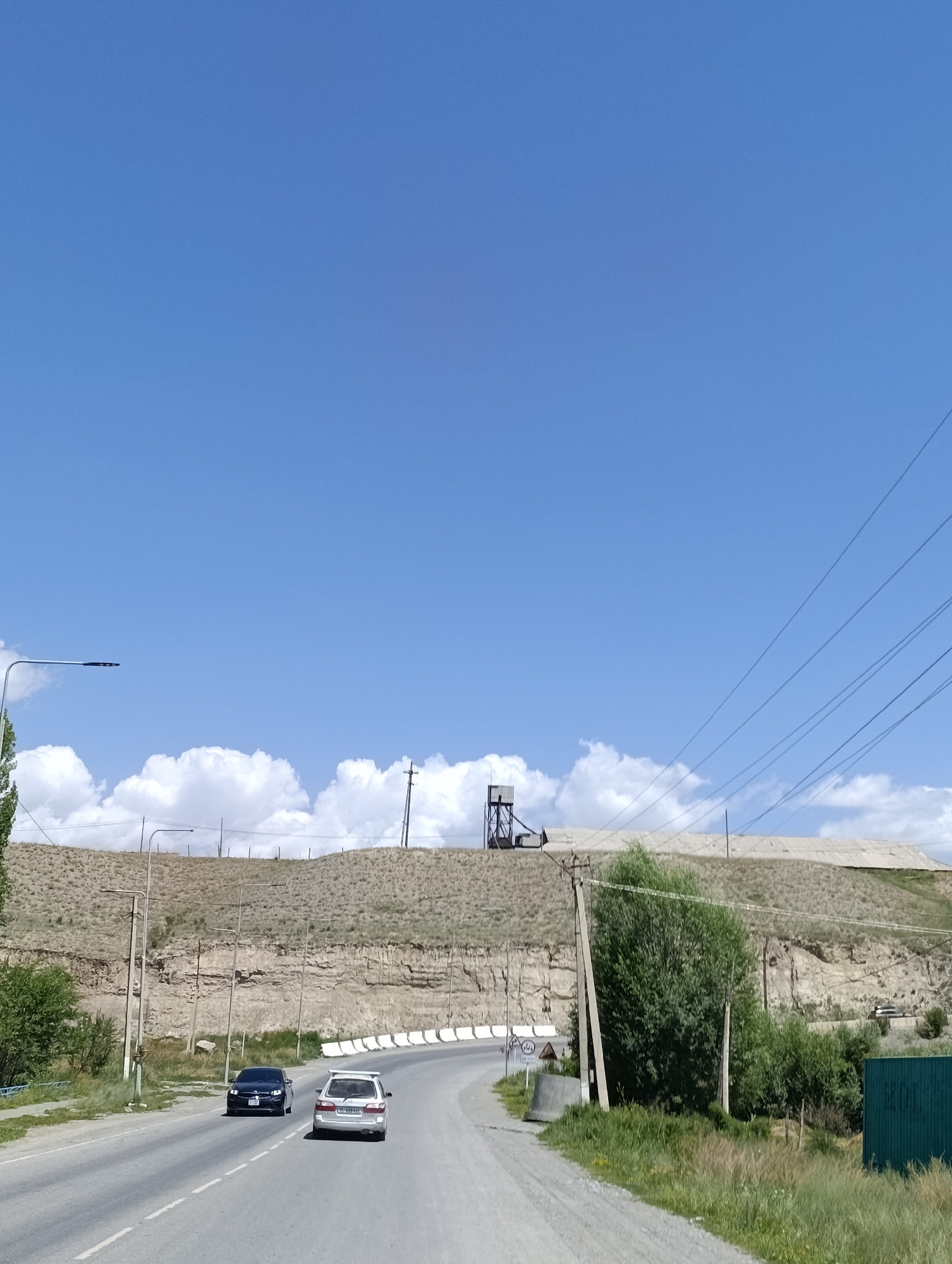 The image shows a view of a road in At-Bashy, Kyrgyzstan, under a clear blue sky with scattered white clouds. The road curves to the right and is lined with grassy verges and trees. In the foreground, two cars are driving on the road: a dark sedan and a white SUV. Beyond the road, a steep, rocky embankment rises, dotted with sparse vegetation and several utility poles. On top of the embankment, there is a large industrial structure with a metal tower, and a long, flat building with a corrugated roof. The scene appears to be a rural or semi-industrial area.