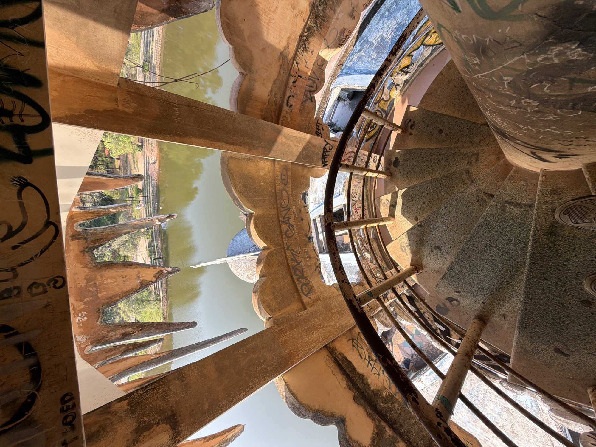 An overhead shot reveals a weathered spiral staircase in the city of Thủy Xuân Ward, Vietnam. The staircase's center column is heavily marked with graffiti. The treads of the staircase are textured. A rusty metal railing runs along the inner edge of the staircase. Outside the staircase, a moat-like body of water is visible, bordered by architectural features resembling large teeth or spikes. Graffiti is also visible on these exterior walls. The lighting is bright, casting stark shadows across the scene. The overall impression is one of decay and neglect within an unusual architectural setting.