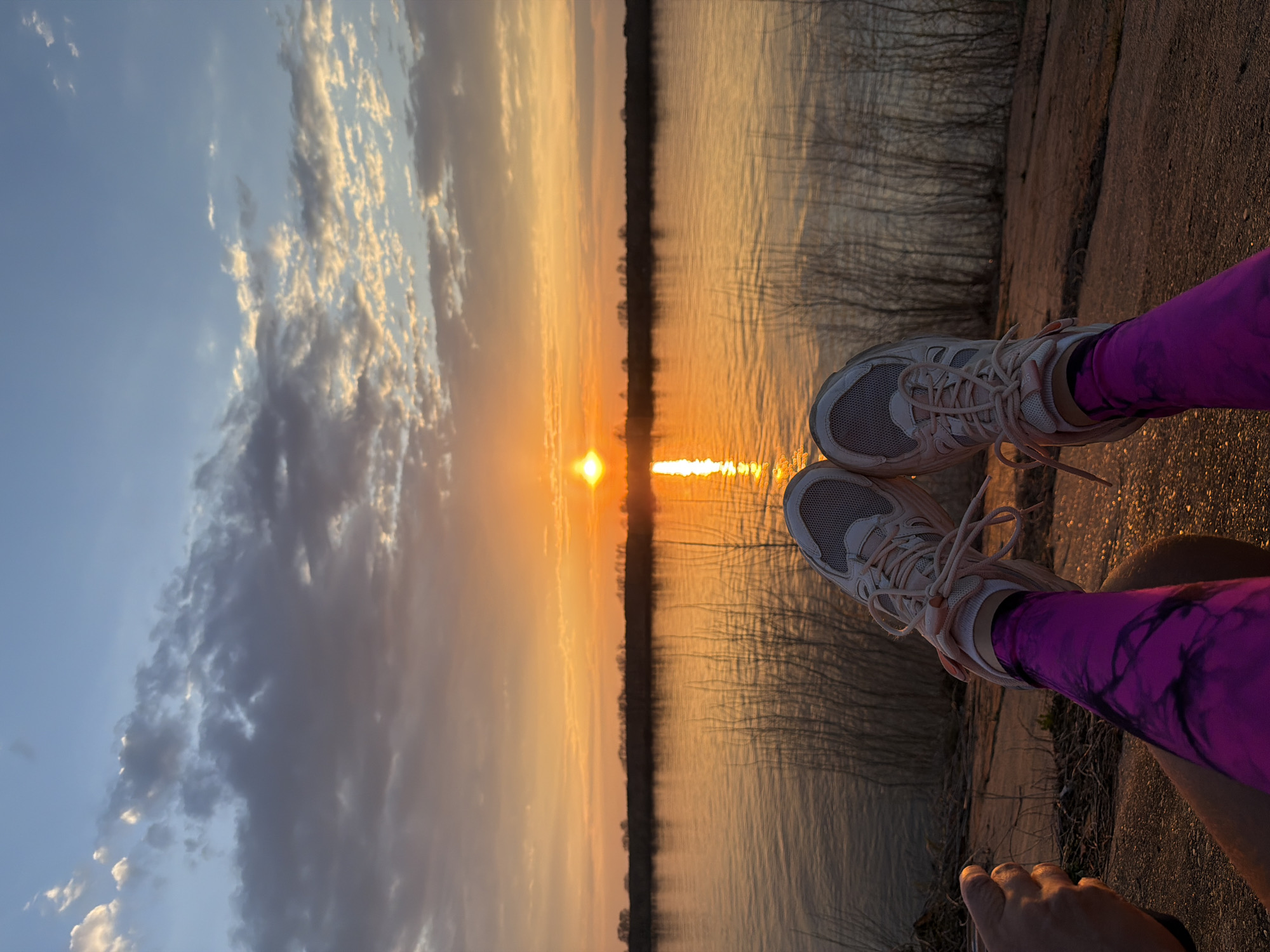 A sunset over a body of water is viewed from a bank, likely in Chaikovsky, Russia. The sky is partially cloudy, transitioning from blue to orange and yellow hues near the horizon where the sun reflects brightly on the water. The opposite bank appears to be wooded with bare trees silhouetted against the sky. A person's legs wearing running shoes and purple leggings are in the foreground, positioned as if sitting or resting by the water's edge. Their hand is visible in the bottom corner. The bank on which the person sits is made of concrete or stone.