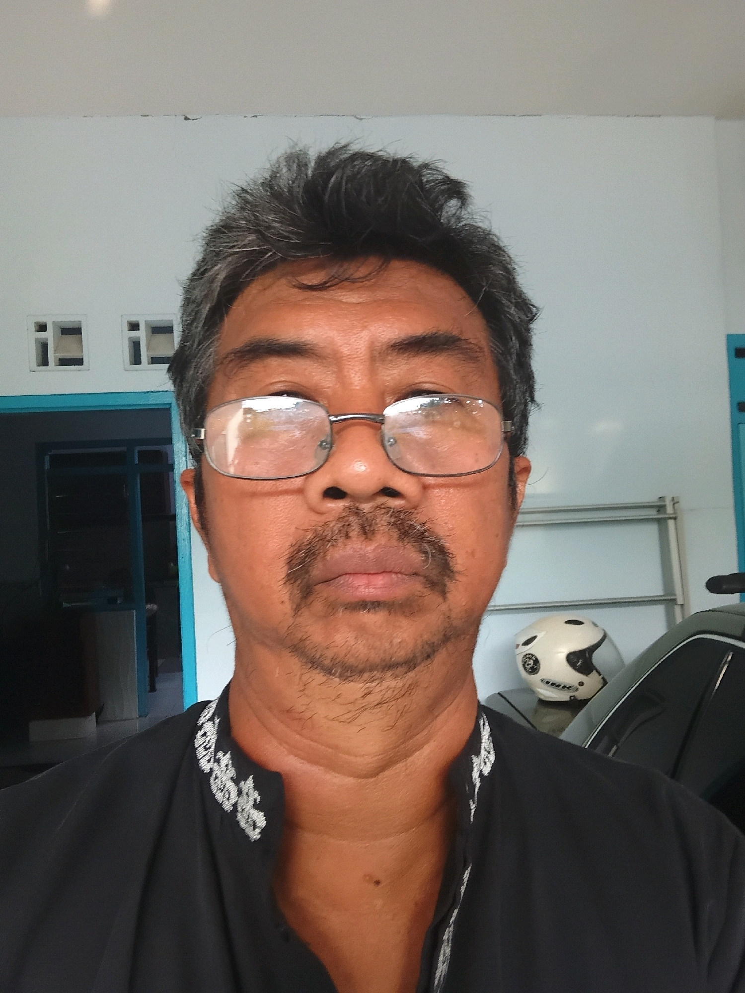 A medium shot photo shows a middle-aged Indonesian man with graying hair, a mustache, and a goatee. He is wearing glasses and a black collared shirt with white embroidered detailing on the collar. He is standing in what appears to be a residential area in Tanggul Wetan, Indonesia. In the background, there is a light blue wall, a doorway with a blue frame, and a white shelf unit. To the right of the man, there is a motorcycle helmet and a portion of a dark-colored vehicle. The lighting suggests it is daytime.