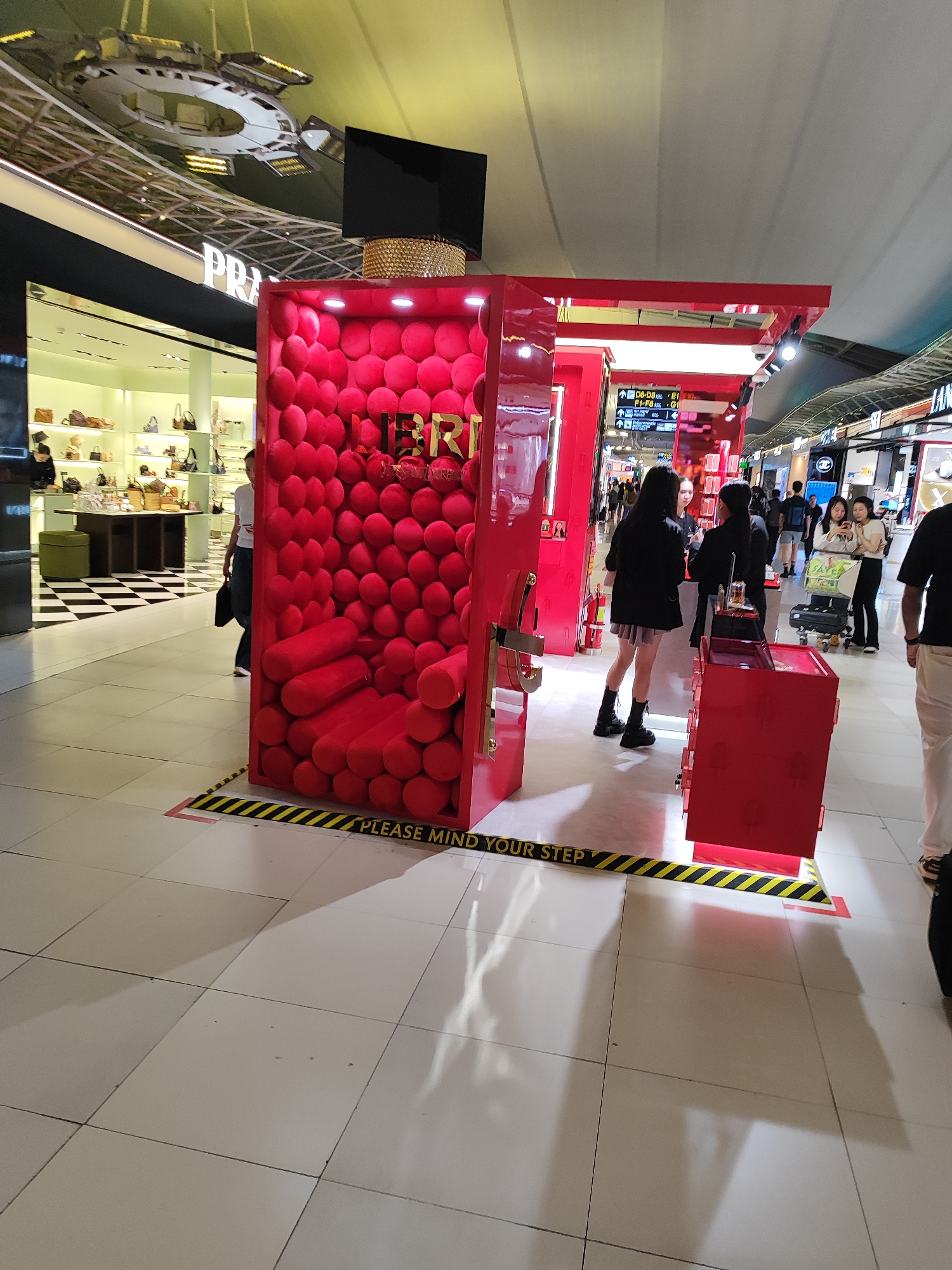 This image shows a promotional display for a brand, identified by the word "IBRI" visible on the structure. The display is designed as a large red rectangle filled with red cushion-like spheres, and a red cart-like object sits nearby. The display is surrounded by yellow and black caution tape that reads "PLEASE MIND YOUR STEP." Several people are present, including two women standing near the display, and other individuals are visible in the background, suggesting a public space, possibly an airport. The scene is indoors with bright overhead lighting. A luxury store, Prada, is visible in the background. The location is Nong Prue Subdistrict, Thailand, based on the context.