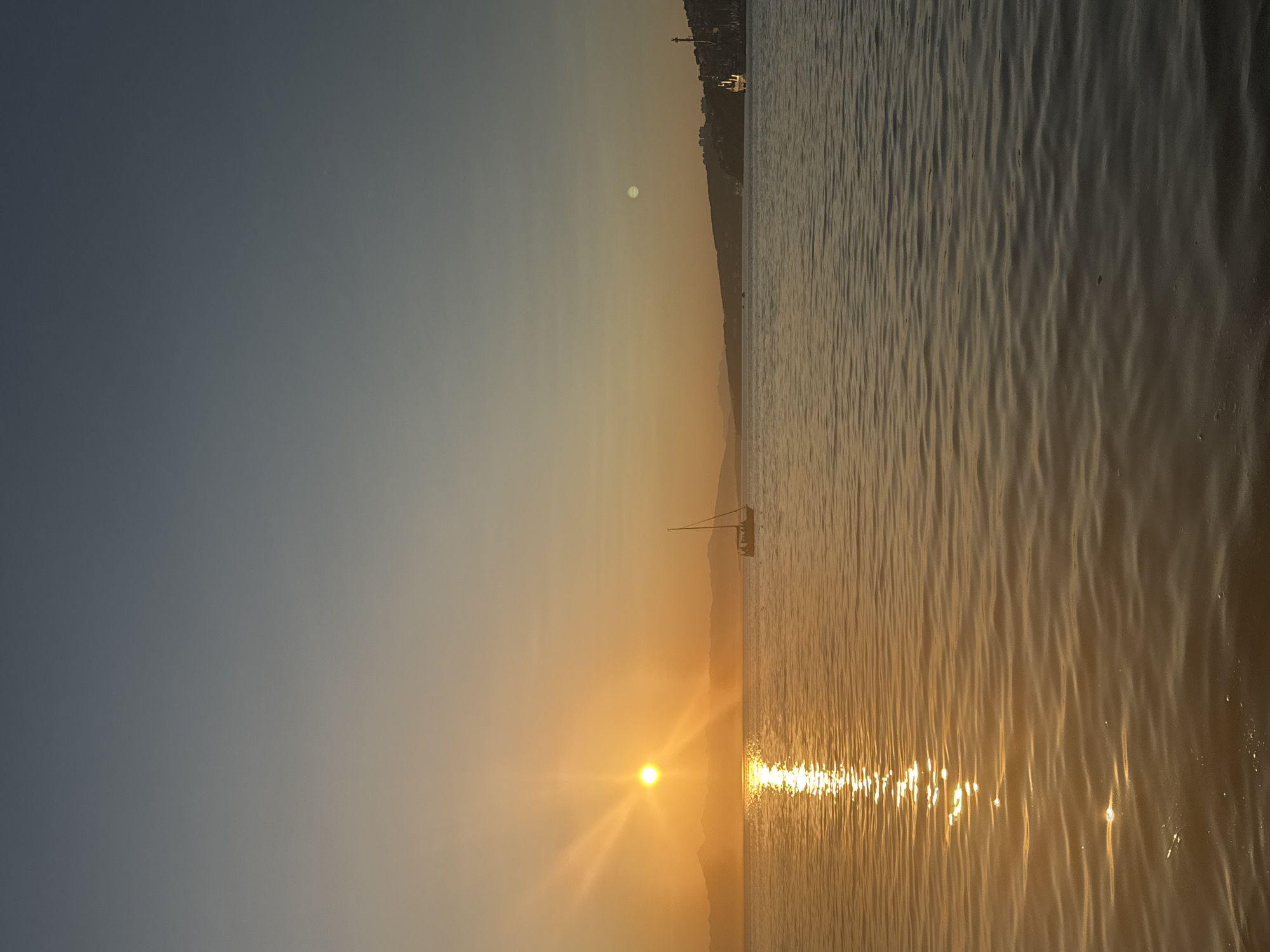 An early morning or late afternoon view of Avacha Bay in Petropavlovsk-Kamchatsky, Russia. The sun, low in the sky and casting a warm orange glow, is visible on the left side of the frame, with its rays stretching across the water. The water itself is calm, with gentle ripples reflecting the sunlight. A sailboat with several people on board is positioned in the mid-ground, to the right of the sun. In the distance, a hilly coastline is visible, with some buildings and structures on the right side of the bay, including what appears to be a port or industrial area with a large ship. The sky transitions from a bright orange near the sun to a muted blue at the top of the frame, indicating a transition from daylight to twilight or vice-versa. A faint, pale moon is visible in the upper right quadrant of the sky.