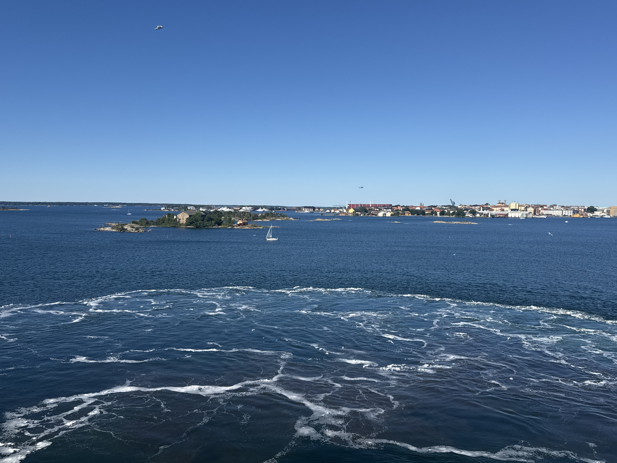 The image shows a wide view of a body of water, likely a harbor or archipelago, with a city skyline in the distance. The foreground is dominated by the turbulent wake of a boat, characterized by frothy white lines and churned water, indicating movement. Beyond this, the water is a deep blue, with smaller ripples. In the middle ground, several small islands are visible, some with buildings and vegetation. A white sailboat is present on the water. The background features a city with a varied architecture, including red-roofed buildings, taller structures, and cranes, suggesting an urban port area. The sky is a clear, bright blue with a single white bird flying high. The scene appears to be Karlskrona, Sweden, given the characteristic archipelago environment and urban port structures.